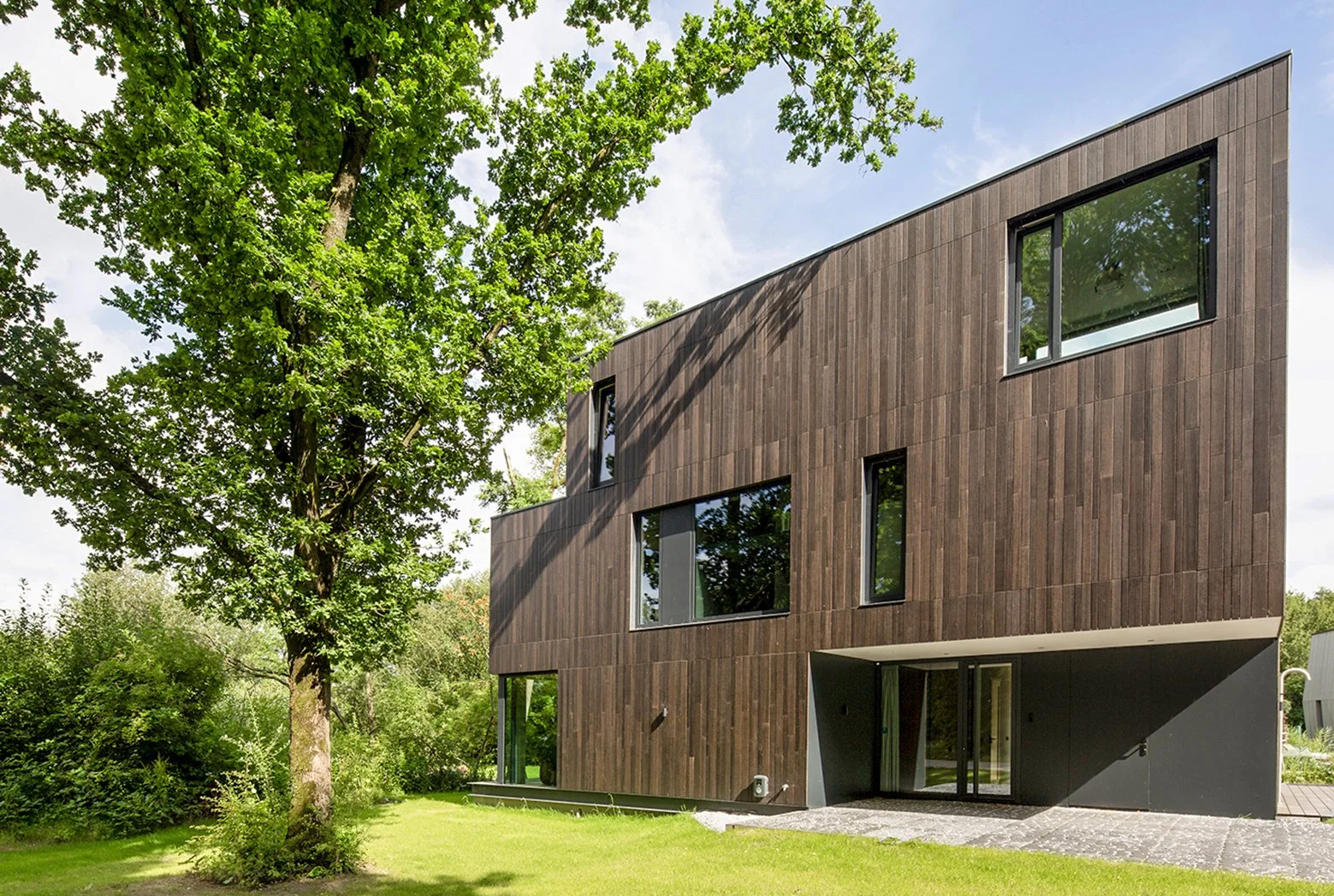 Moderne, drie verdiepingen tellende woning met een houten gevel en grote ramen, omgeven door groene bomen en een gazon.