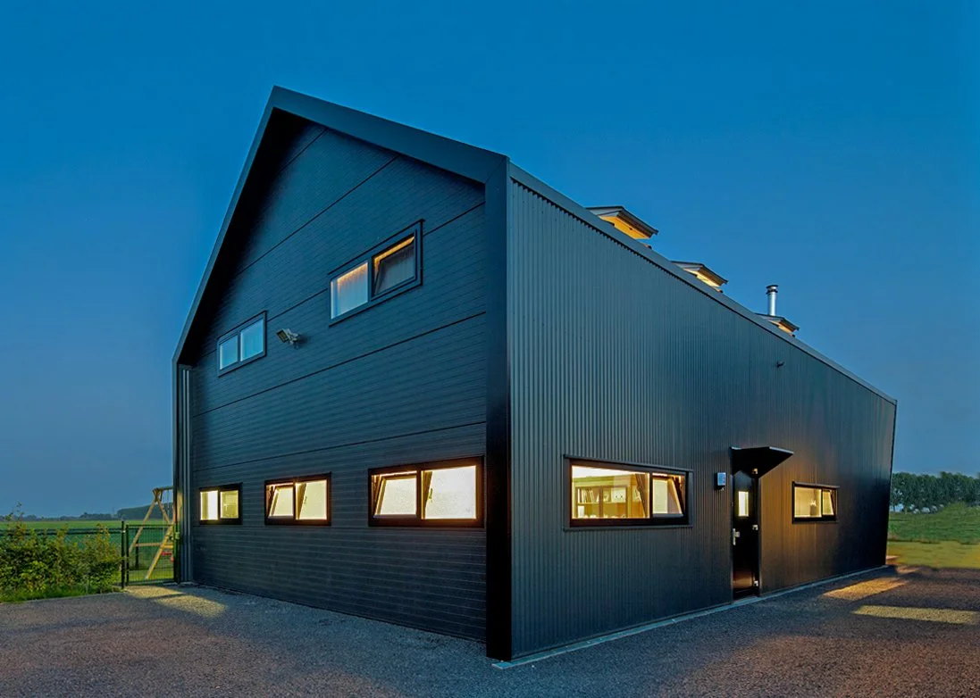 Moderne zwarte woning met schuine dak, grote ramen met verlichting van binnen, avondlucht, groene velden op de achtergrond.