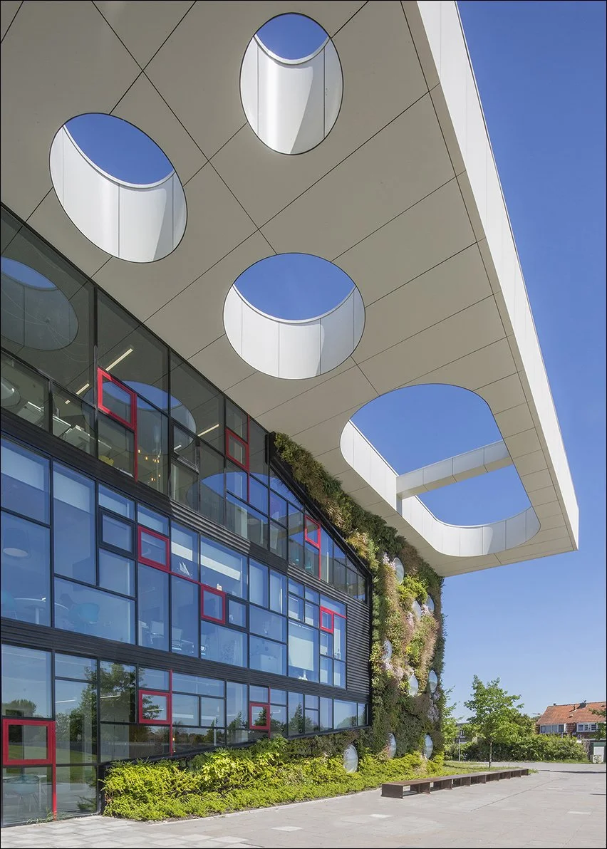 Modern gebouw met grote glasramen en ronde openingen in het plafond, omgeven door groene planten en een open blauwe lucht.