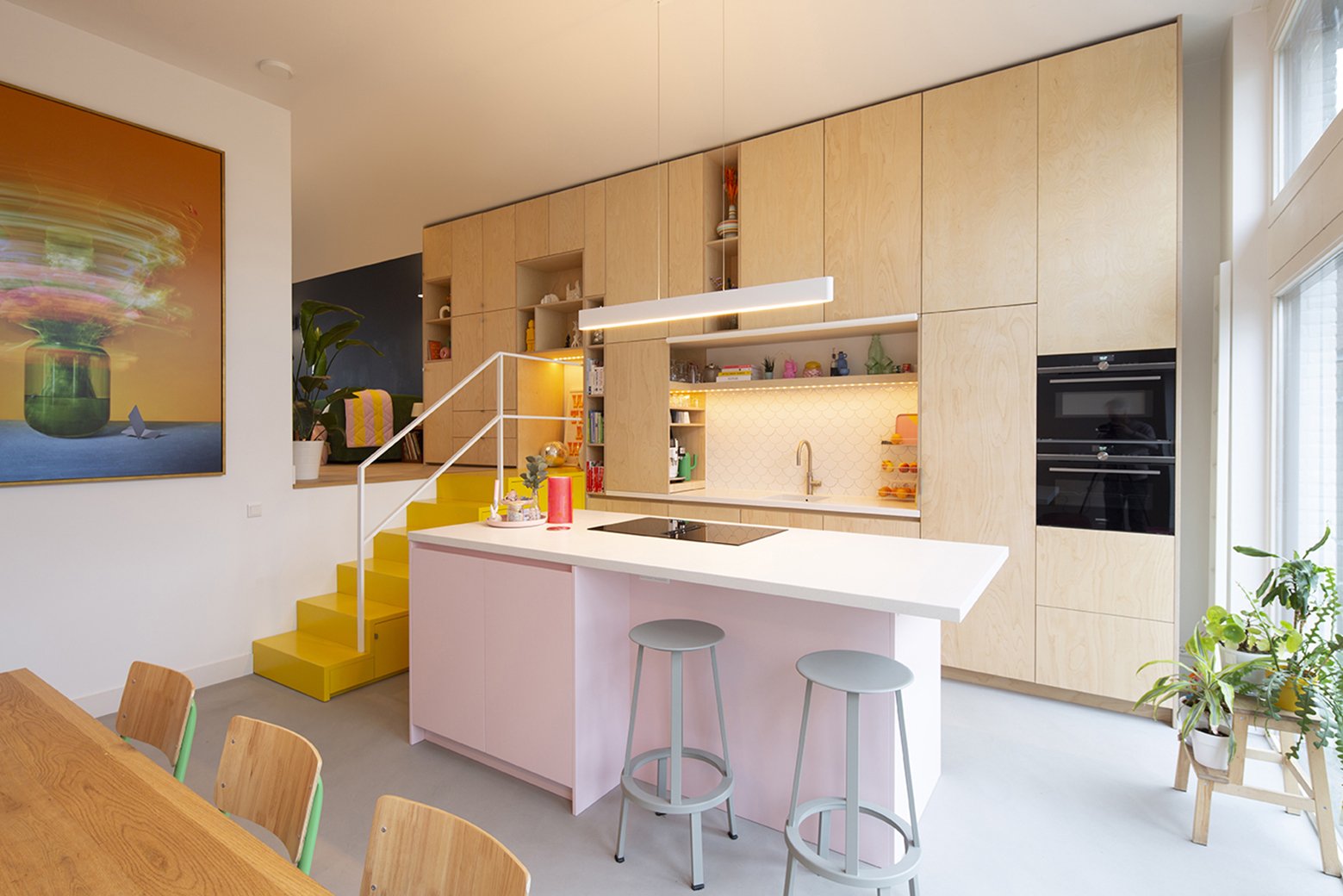Moderne keuken met houten kasten, een wit eiland met roze rand, twee grijze barkrukken, gele trap, planten en ingelichte bovenkastjes.