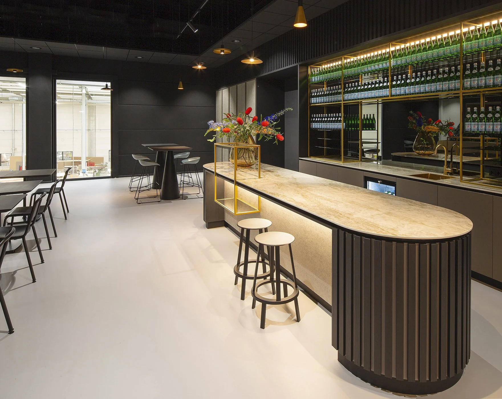 Modern bar met zwarte wanden, een marmeren barblad, gouden accenten, groene flessen op glazen planken en een bloemstuk met kleurrijke bloemen.