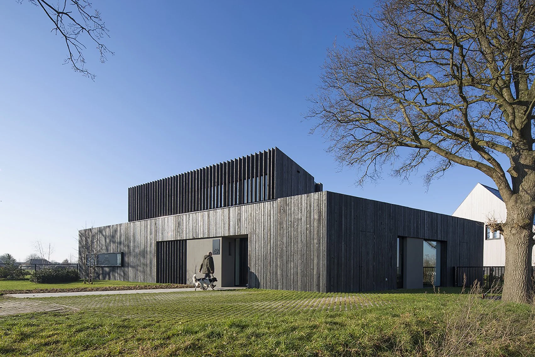 Een modern huis met een houten gevel en zwarte houten latten aan de bovenkant. Een persoon loopt met een hond voor de ingang, onder een grote boom met kale takken onder een heldere blauwe hemel.