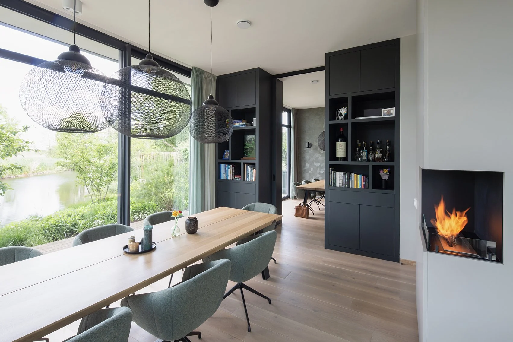 Moderne eetkamer met grote ramen, houten tafel, groene stoelen, zwarte wandrekken, open haard met vlammen, en uitzicht op een tuin met een rivier.