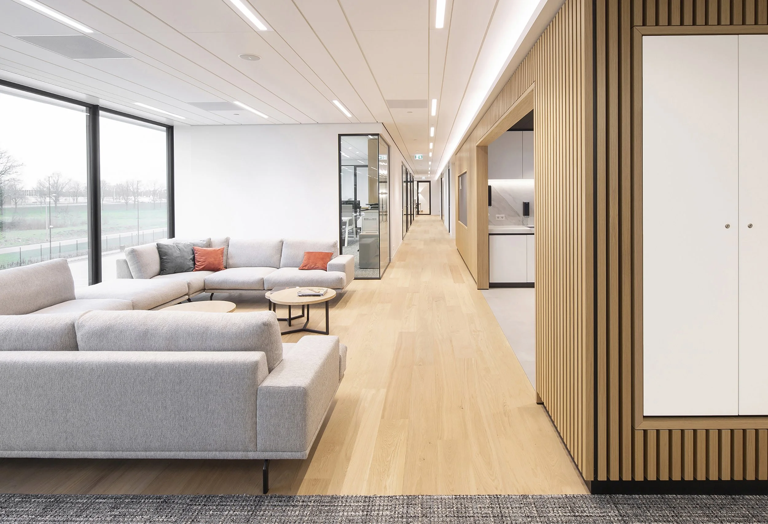 Modern kantoorruimte met grote ramen, een zithoek met lichtgrijze bank, houten vloer, en een wand met verticale houten latten.