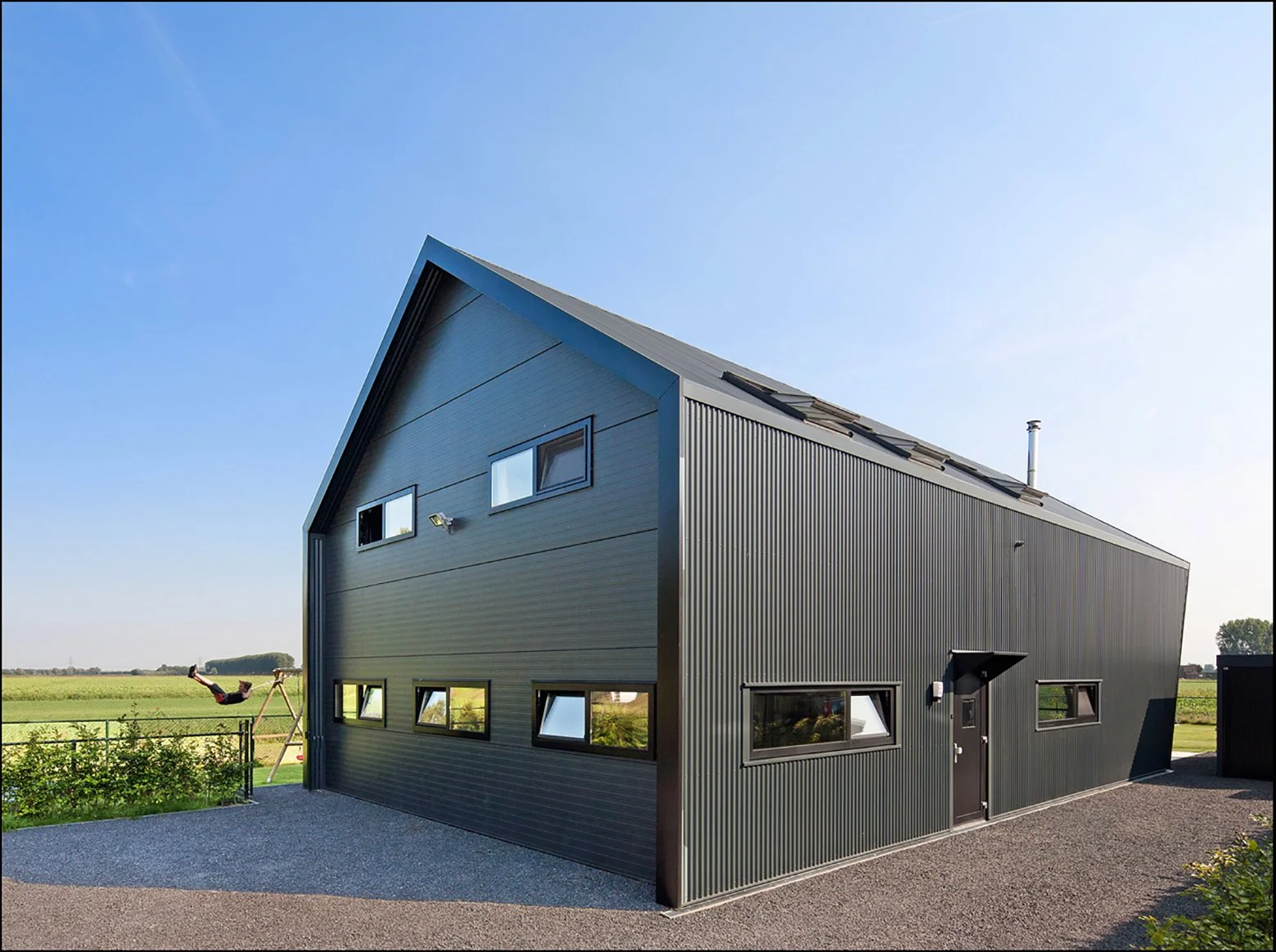 Moderne, zwarte woning met afwijkende gevel en liggend raam, gelegen in een landelijke omgeving met groen gras en blauw hemel.