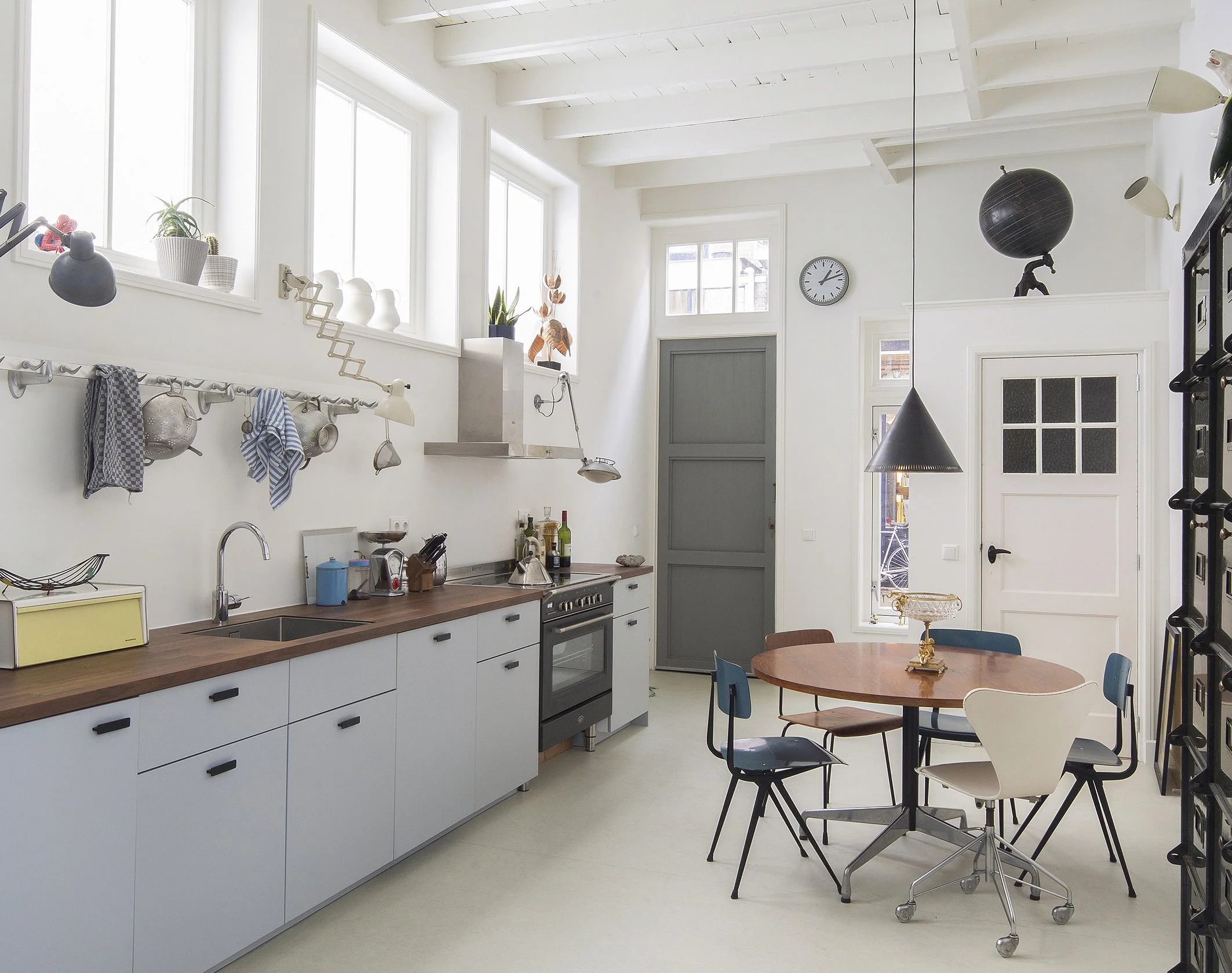 Een ruime, lichte keuken met witte muren en hoge ramen die natuurlijk licht binnenlaten. Er hangen pannen en keukengerei aan de muur, er staan planten op het vensterblad en een klok op de muur. In het midden staat een ronde houten tafel met stoelen e