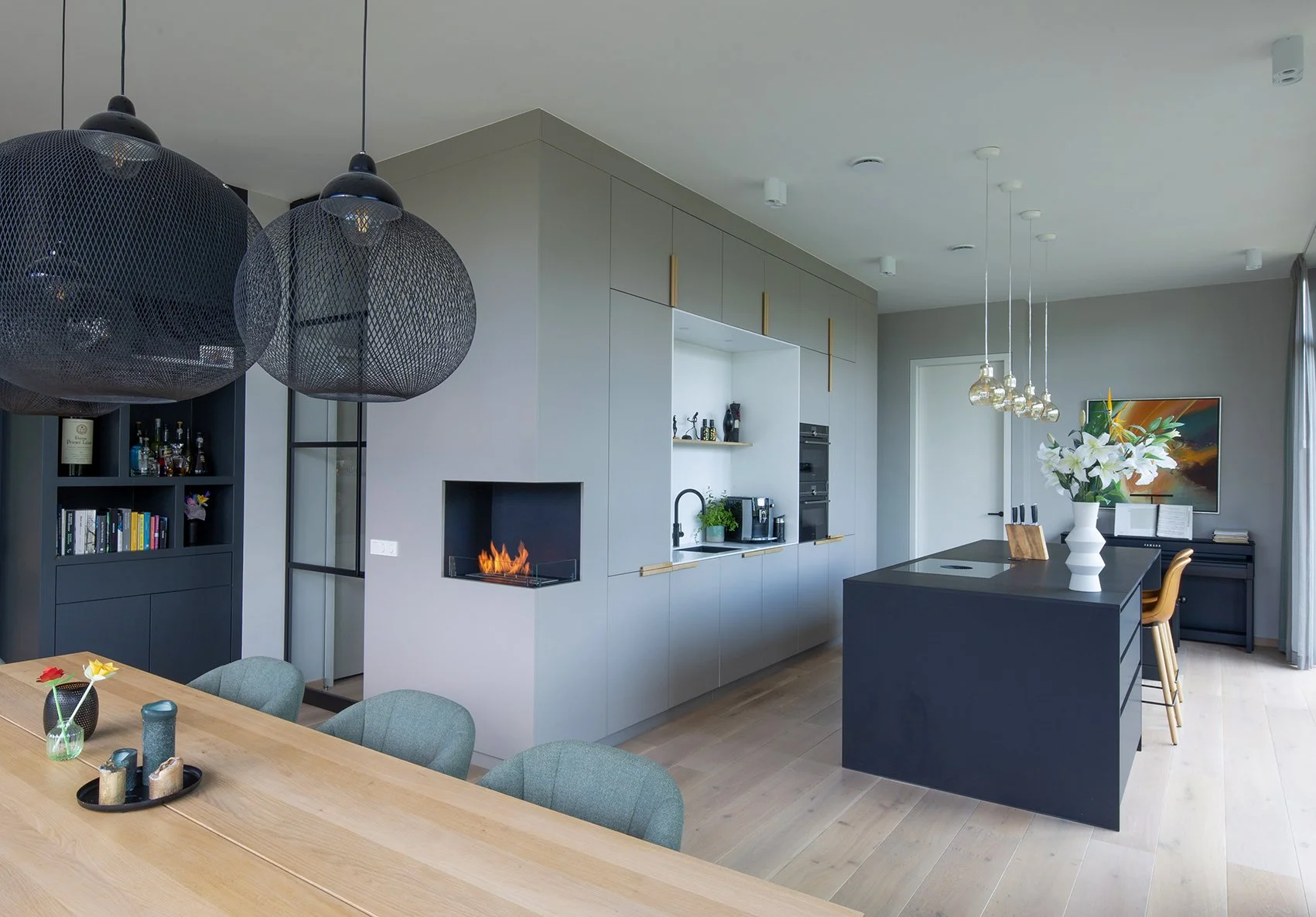 Modern open keuken en eetkamer met lichte kasten, zwart aanrecht en eiland, hanglampen, bloemen, televisie en boeken in een minimalistisch interieur.