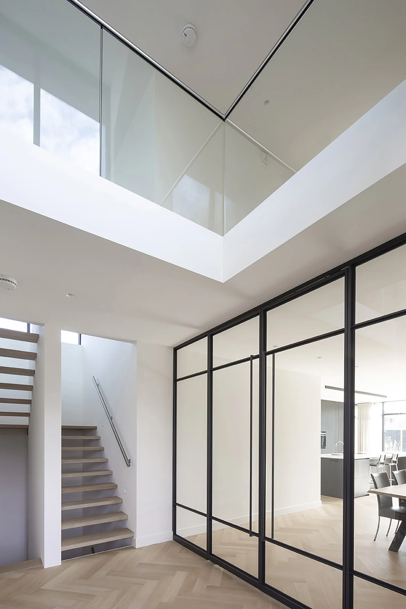 Interieur van een modern huis met een open woonkamer en een trap naar de bovenverdieping, grote glazen wanden en houten vloeren.