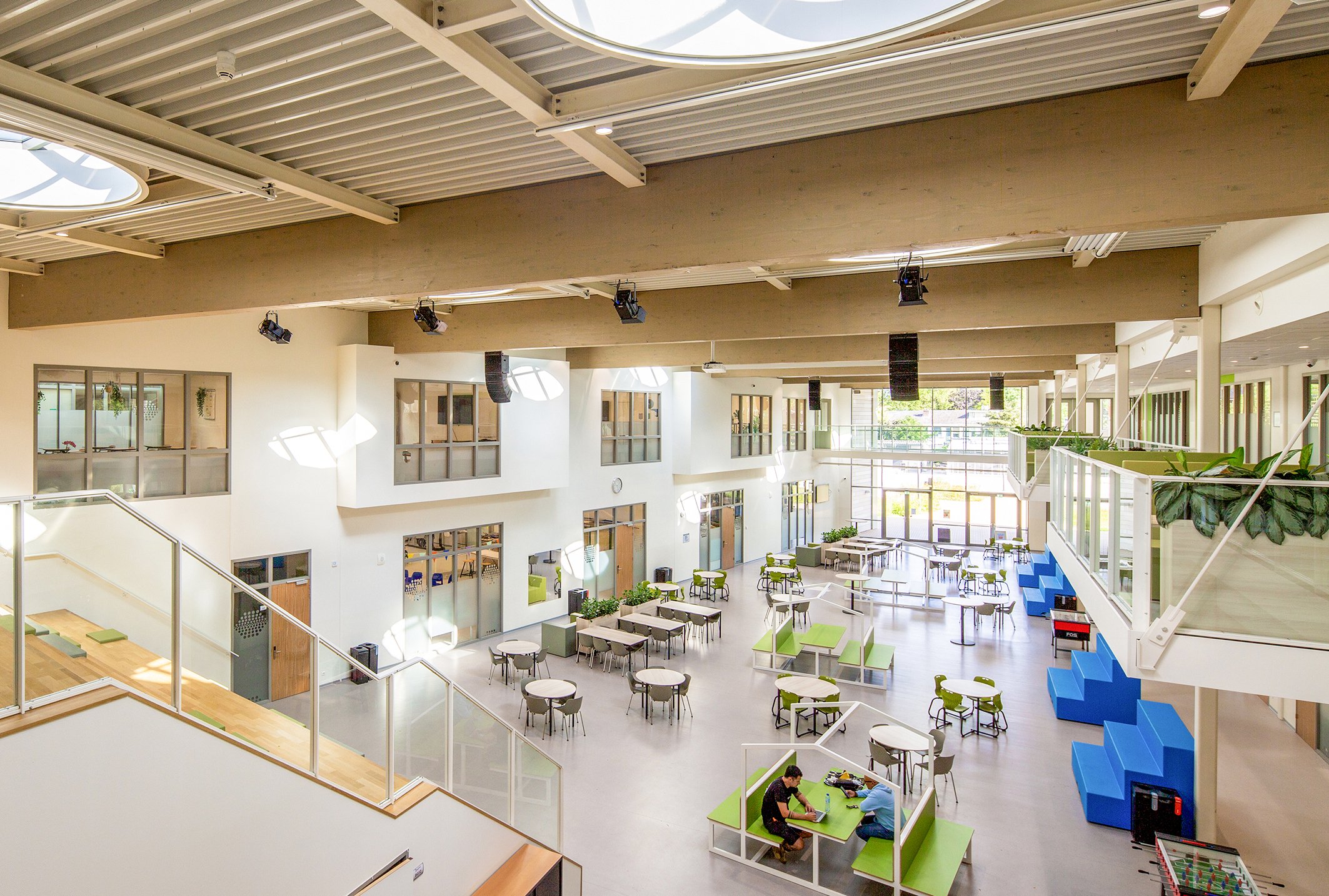Binnenruimte van een modern gebouw met veel ramen, groene decoraties, en verschillende zitplaatsen, waaronder tafels en stoelen. Er hangen speakers, en er is een gezellige sfeer.
