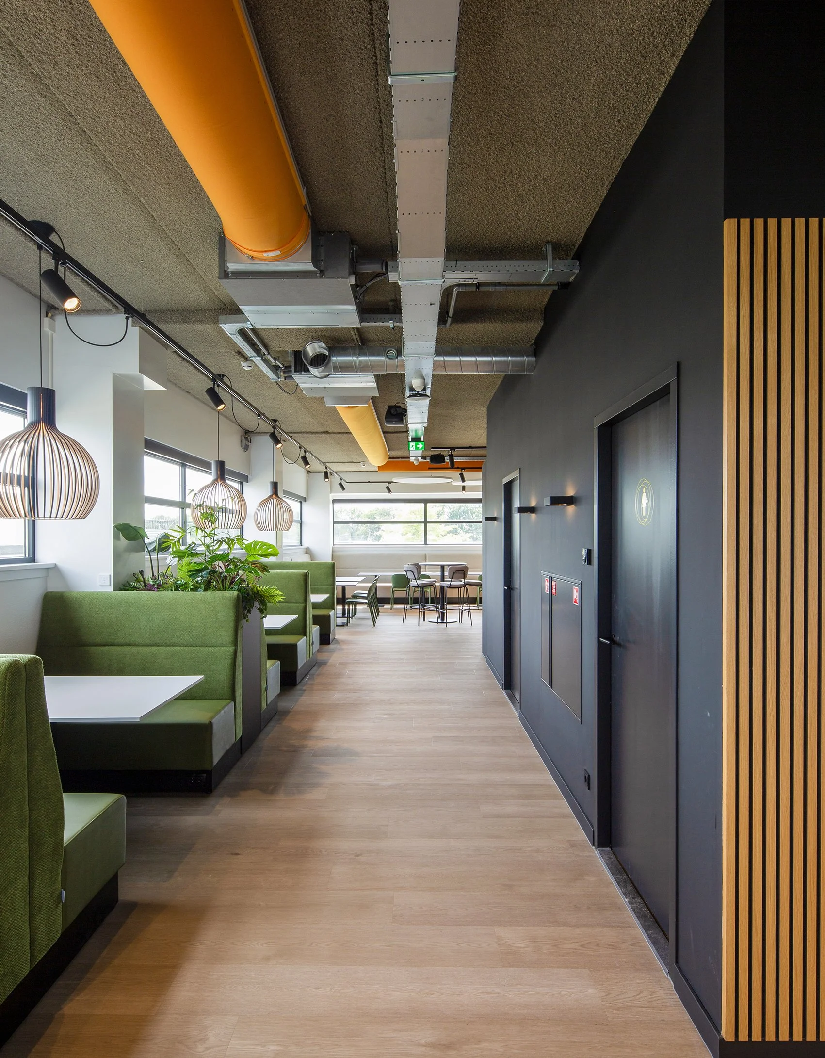 Interieur van een modern kantoor of café met groene stoelen, hanglampen, grote ramen, planten, en een plafond met zichtbare installaties.