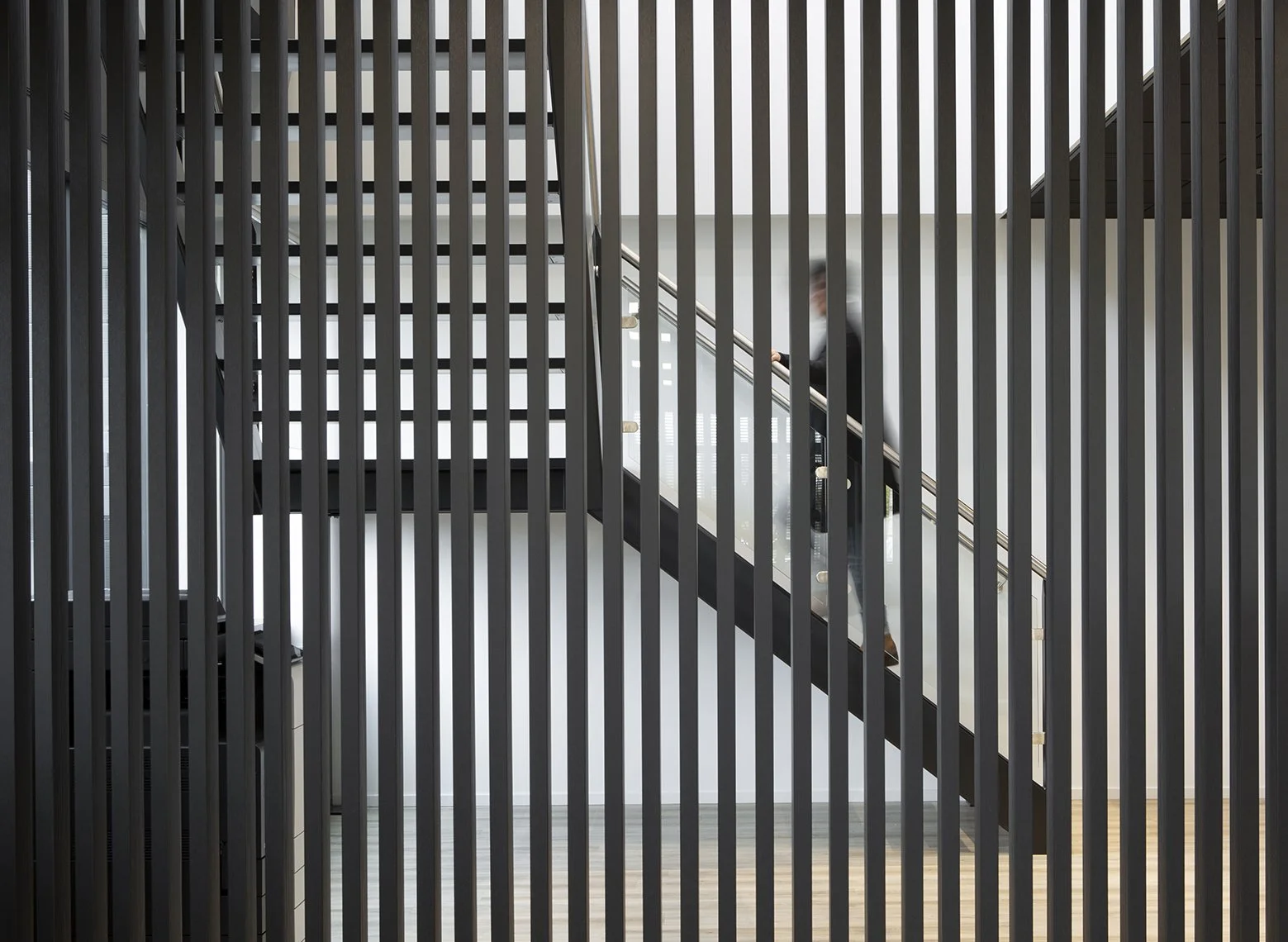 Binnen in een modern gebouw met zwarte en witte muren, zichtbaar door een metalen tralie scheiding, een persoon in een zwart pak die de trap oploopt.