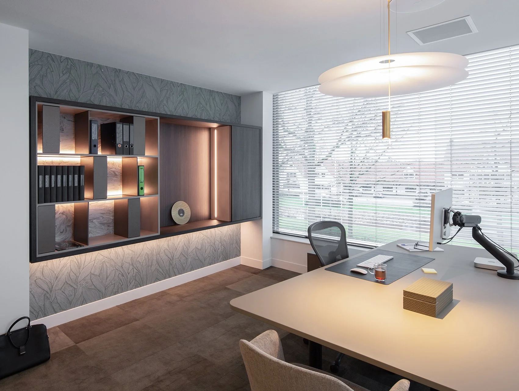 Modern kantoor met bureau, bureaustoel, computer en grote ramen met jaloezieën, een wandrek met mappen en decoratieve verlichting.