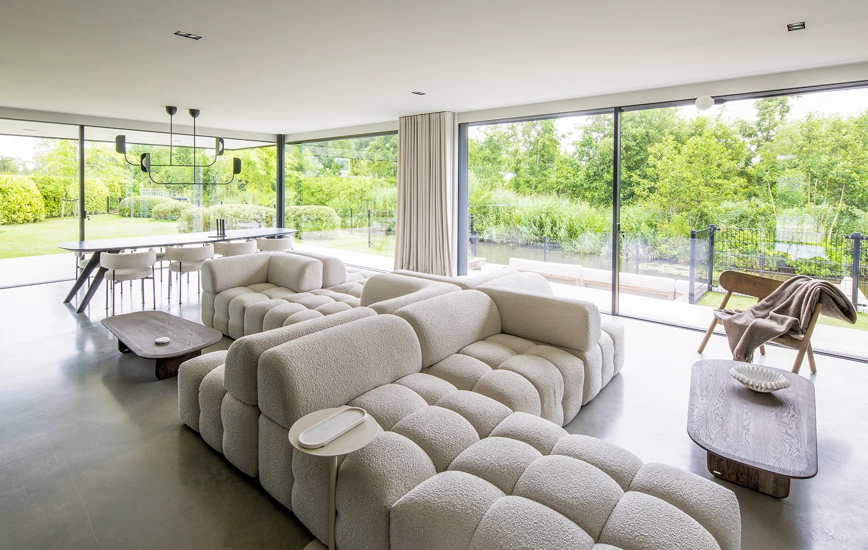 Moderne woonkamer met grote glazen schuifdeuren, witte bank, houten salontafel, en uitzicht op een groene tuin.