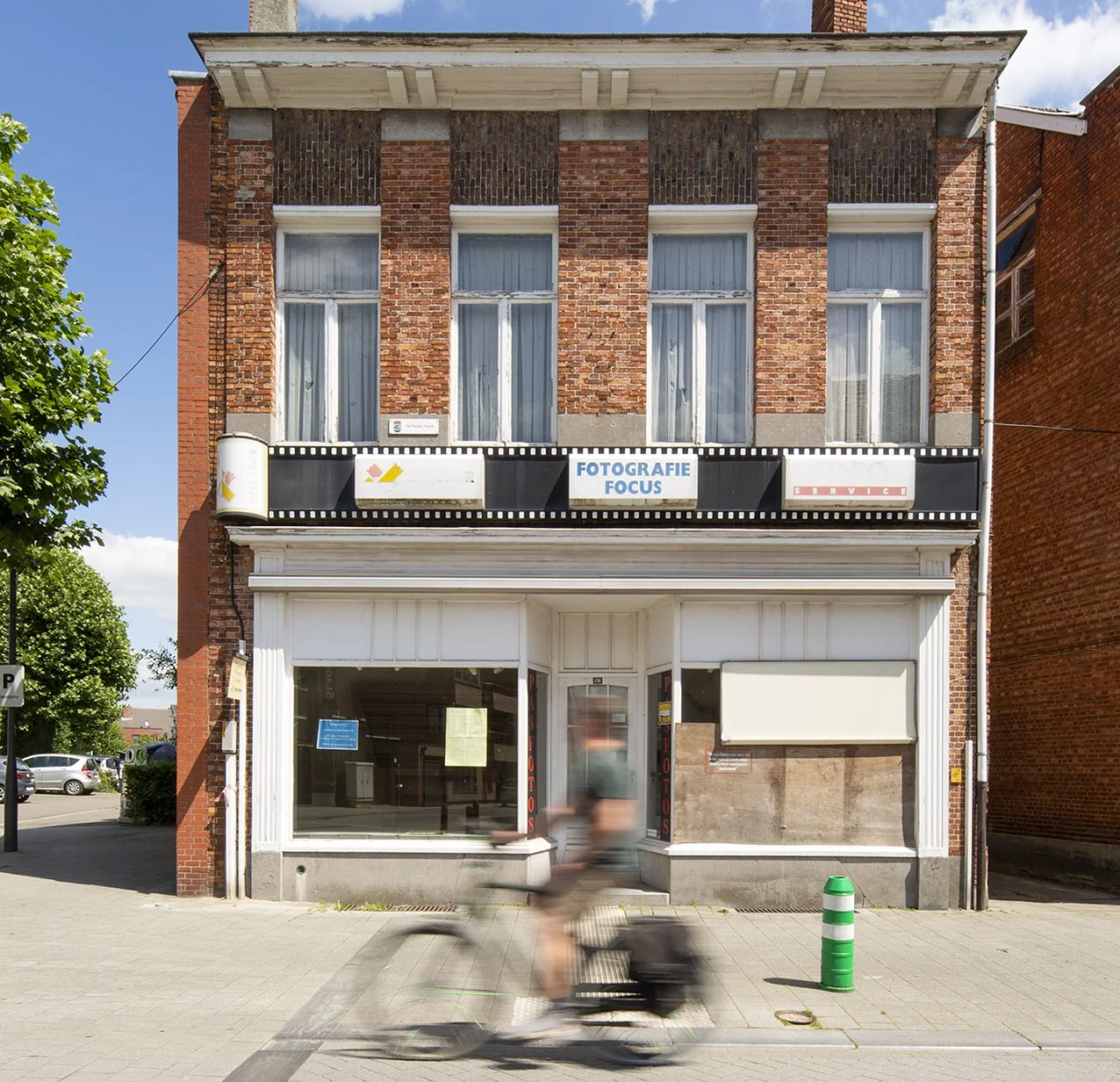 Een oud rode bakstenen gebouw met grote ramen, een fotowinkel met de naam 'Fotografie Focus' en een blurry man op een scooter die voorbijrijdt op een stoep. Buiten een groene en witte verkeerspaal.