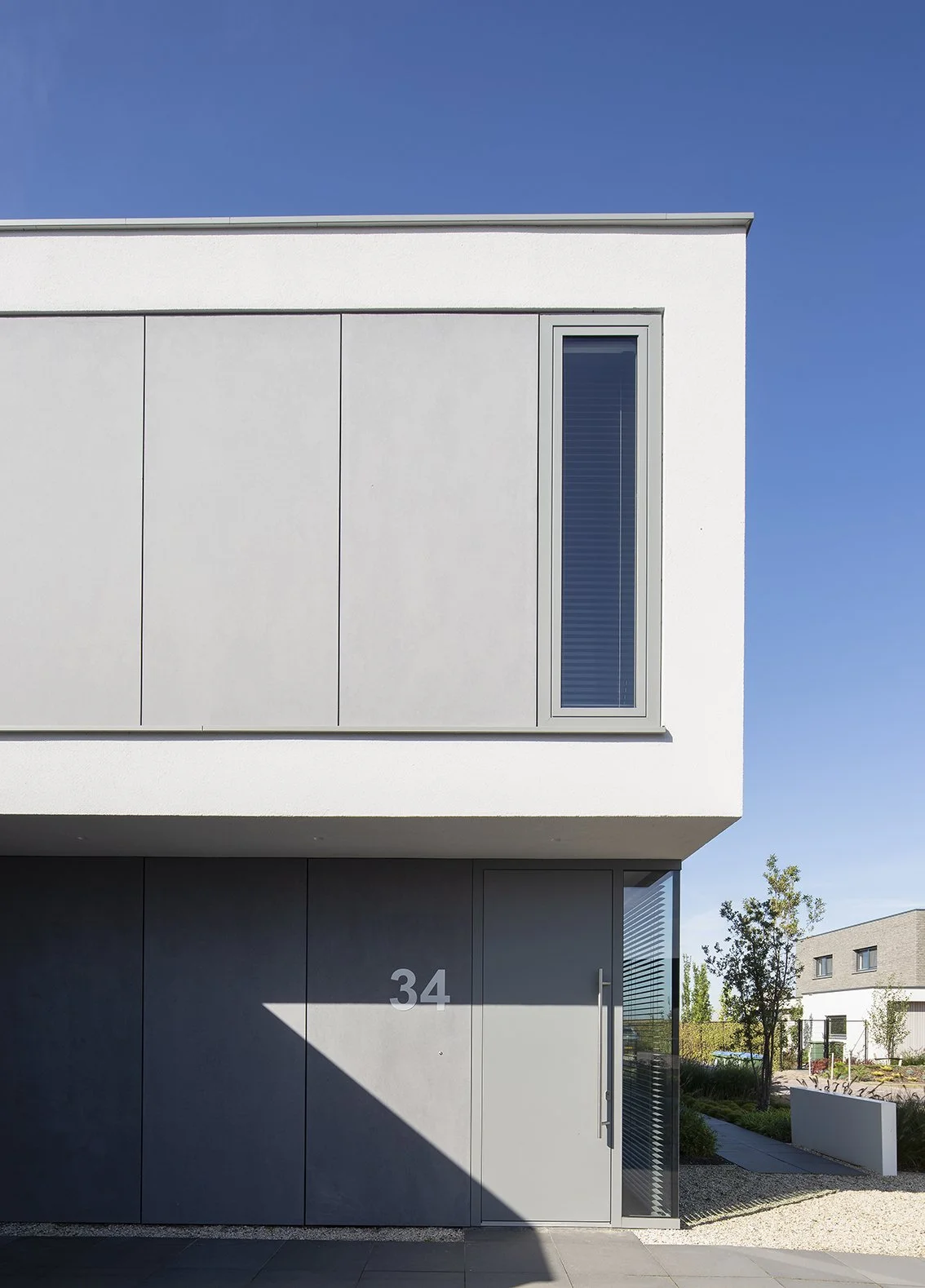 Moderne witte en grijze woning met het nummer 34 op de deur, grote raam en duidelijke architectuur, onder een heldere blauwe hemel.