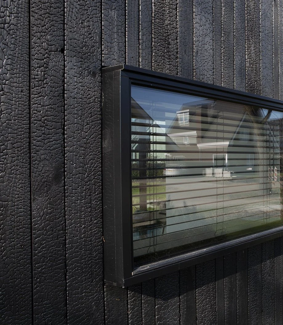 Een zwart geschilderd houten huis met een grote tuin en een raam met horizontale jaloezieën.