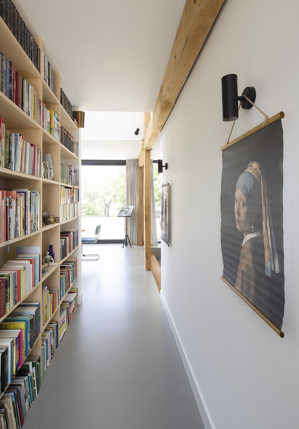 Interieur van een moderne woonkamer met een boekenkast vol boeken aan de linkerkant, een afbeelding van een vrouw rechts, grote ramen met uitzicht op een tuin, en een houten balk over het plafond.
