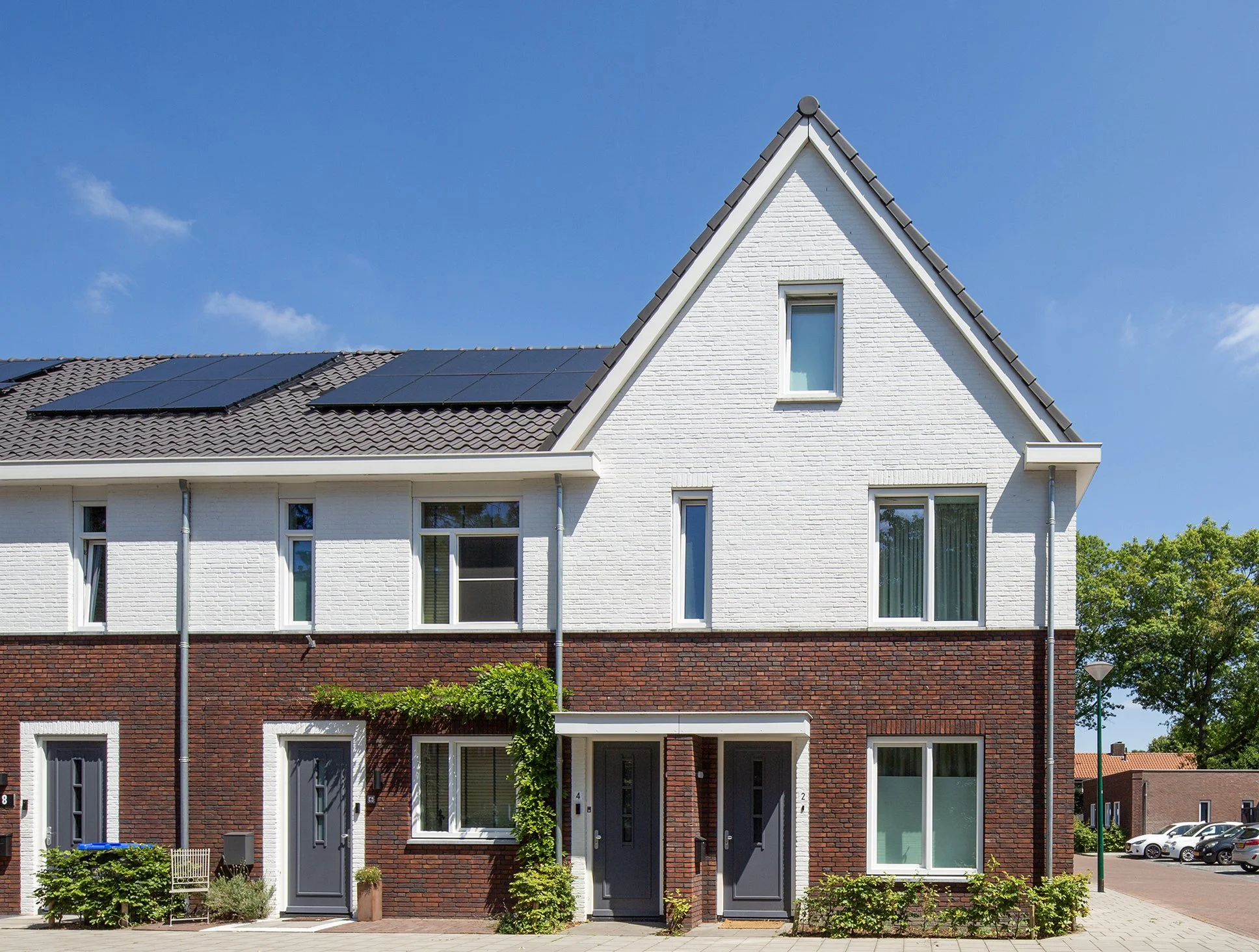 Foto van een moderne witte en rode bakstenen woning met zonnepanelen op het dak en grote ramen, onder een heldere blauwe hemel gefotografeerd door architectuurfotograaf John van Groenedaal.