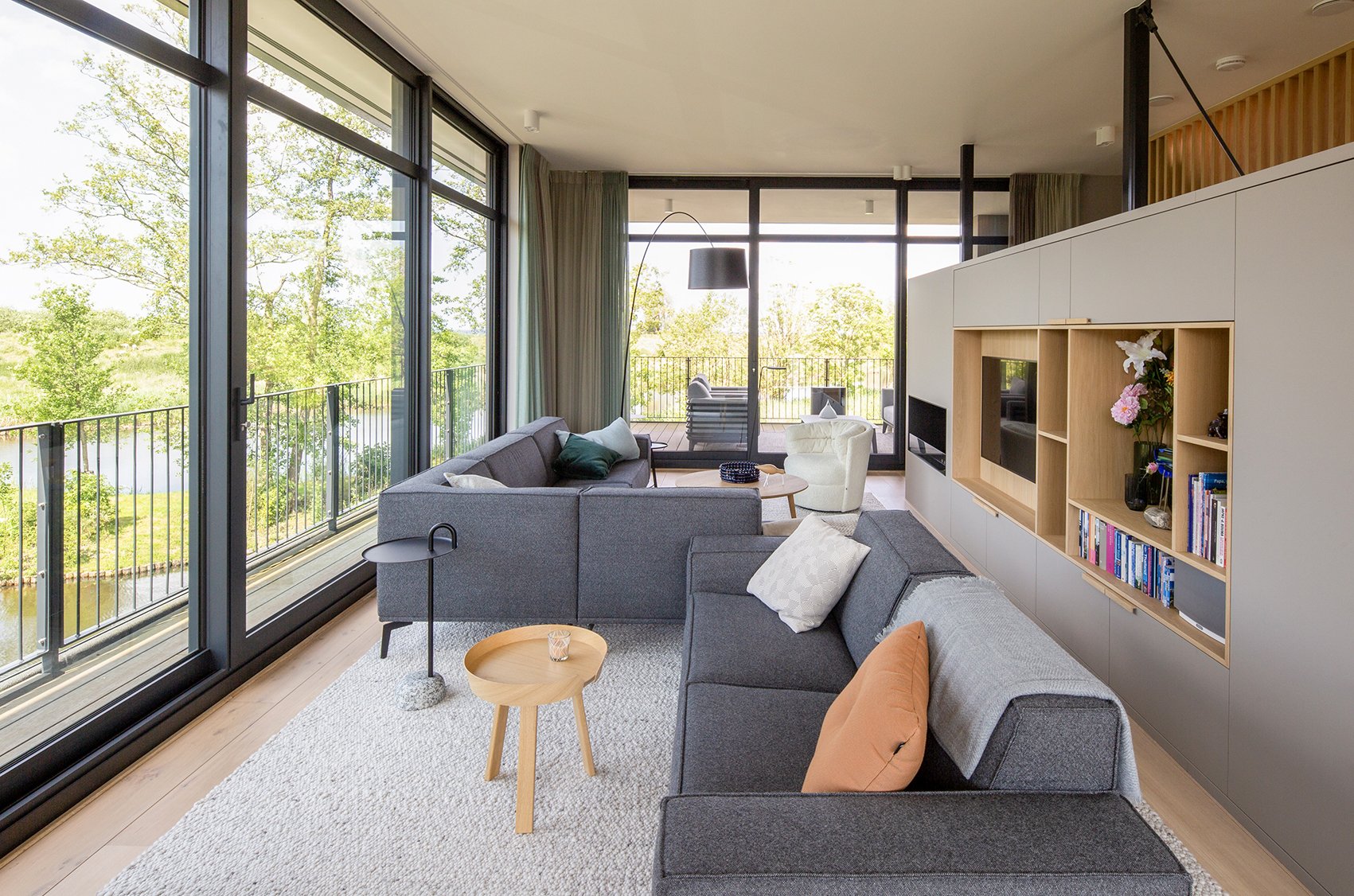 Woonkamer met grote ramen die uitkijken op een groene tuin, inclusief bank, fauteuils, salontafels en een ingebouwde boekenplank met decoraties en boeken.
