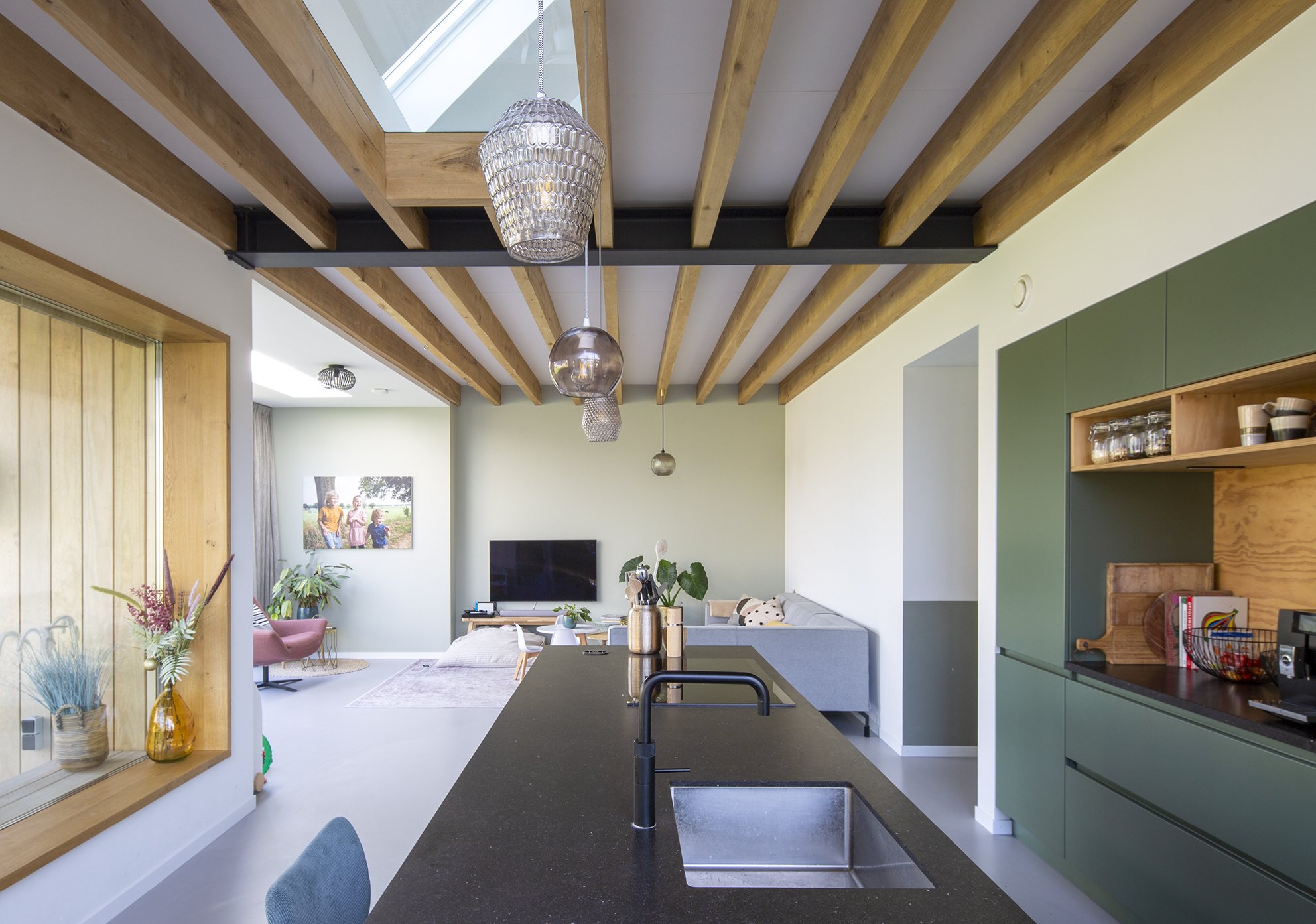Open keuken met groen kasten en een zwart werkblad, hanglampen boven de eettafel, woonkamer met tv en zitplaatsen, planten en decoraties, plafond met houten balken en een dakraam.