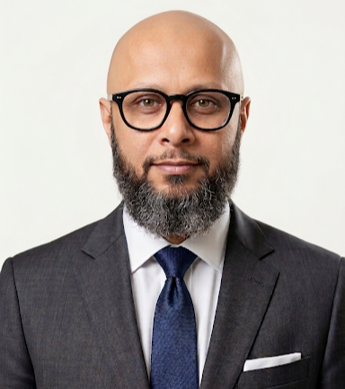 Headshot of a bald man with glasses, a beard, wearing a dark suit, white shirt, and blue tie against a plain white background.