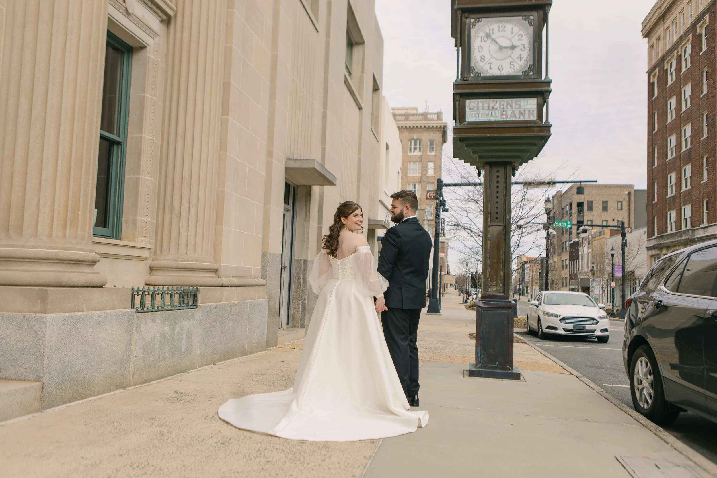 A bride and groom holding hands and looking at each other on a city sidewalk, with a large clock and historic buildings in the background.