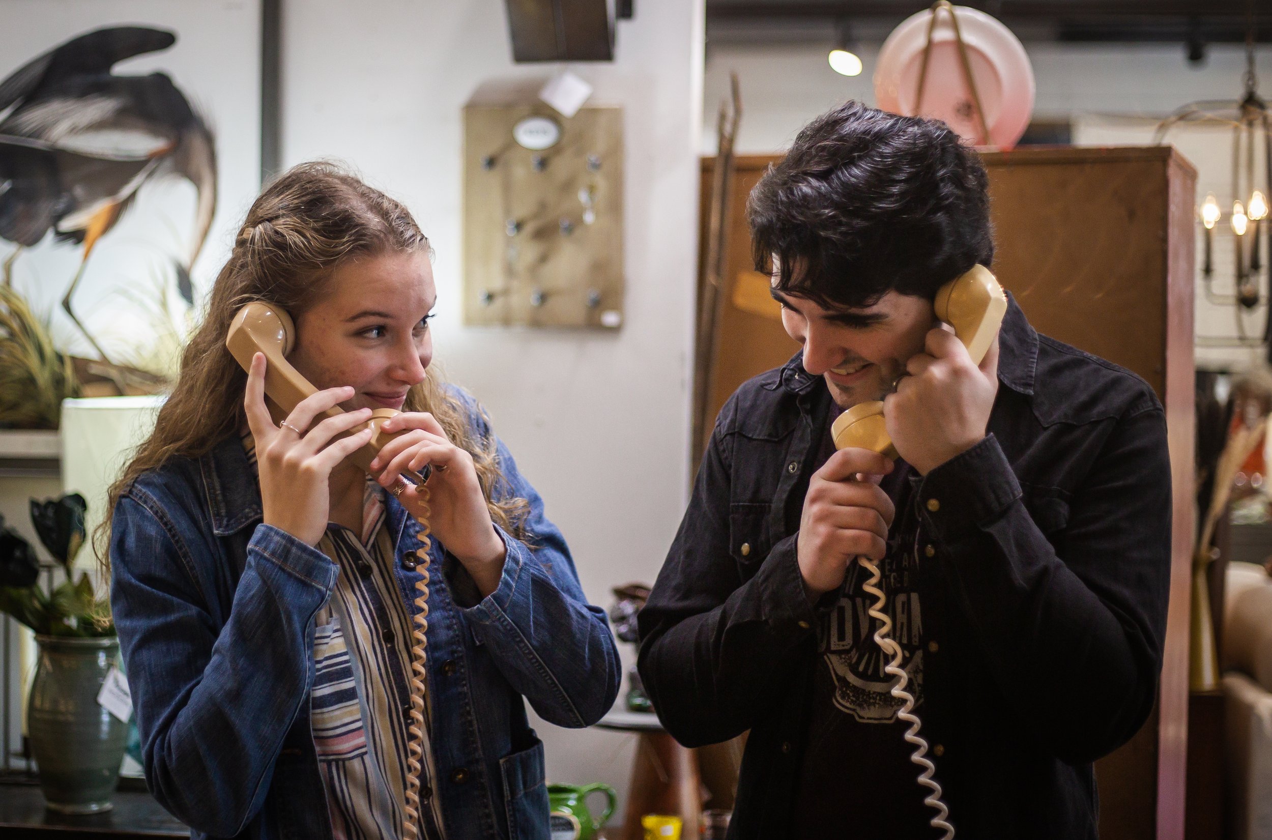 Two young people, a woman with curly hair and a man with dark hair, are talking on vintage beige rotary phones in a cozy, eclectic room.
