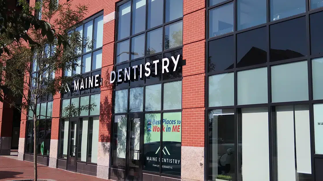 Exterior view of a multi-story building with large glass windows and a red brick facade, housing Maine Dentistry, a family cosmetic dental office.
