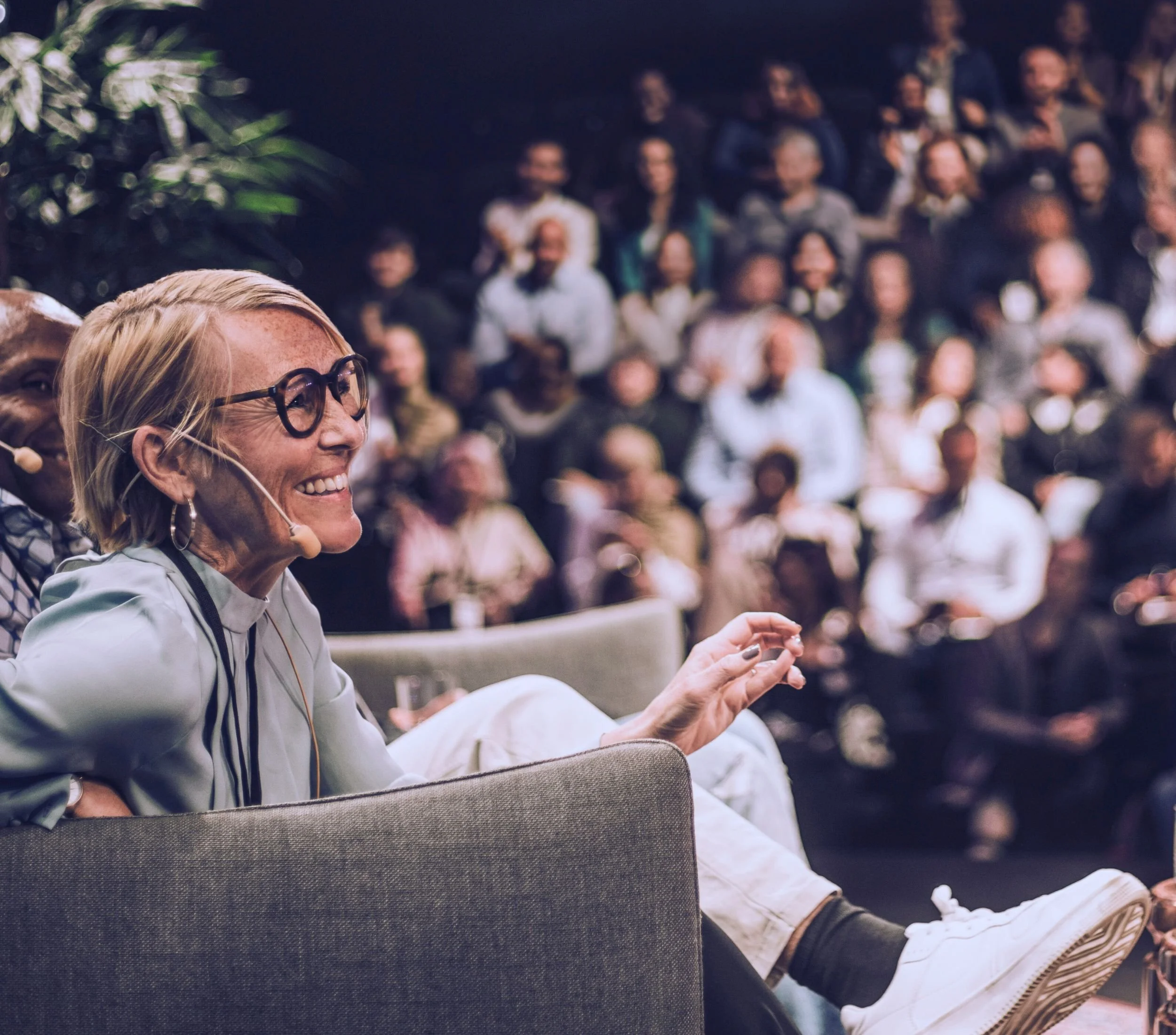 Une femme portant des lunettes et des boucles d'oreilles, souriante et gesticulant, assise sur un canapé lors d'une conférence ou d'un événement, avec un public en arrière-plan.
