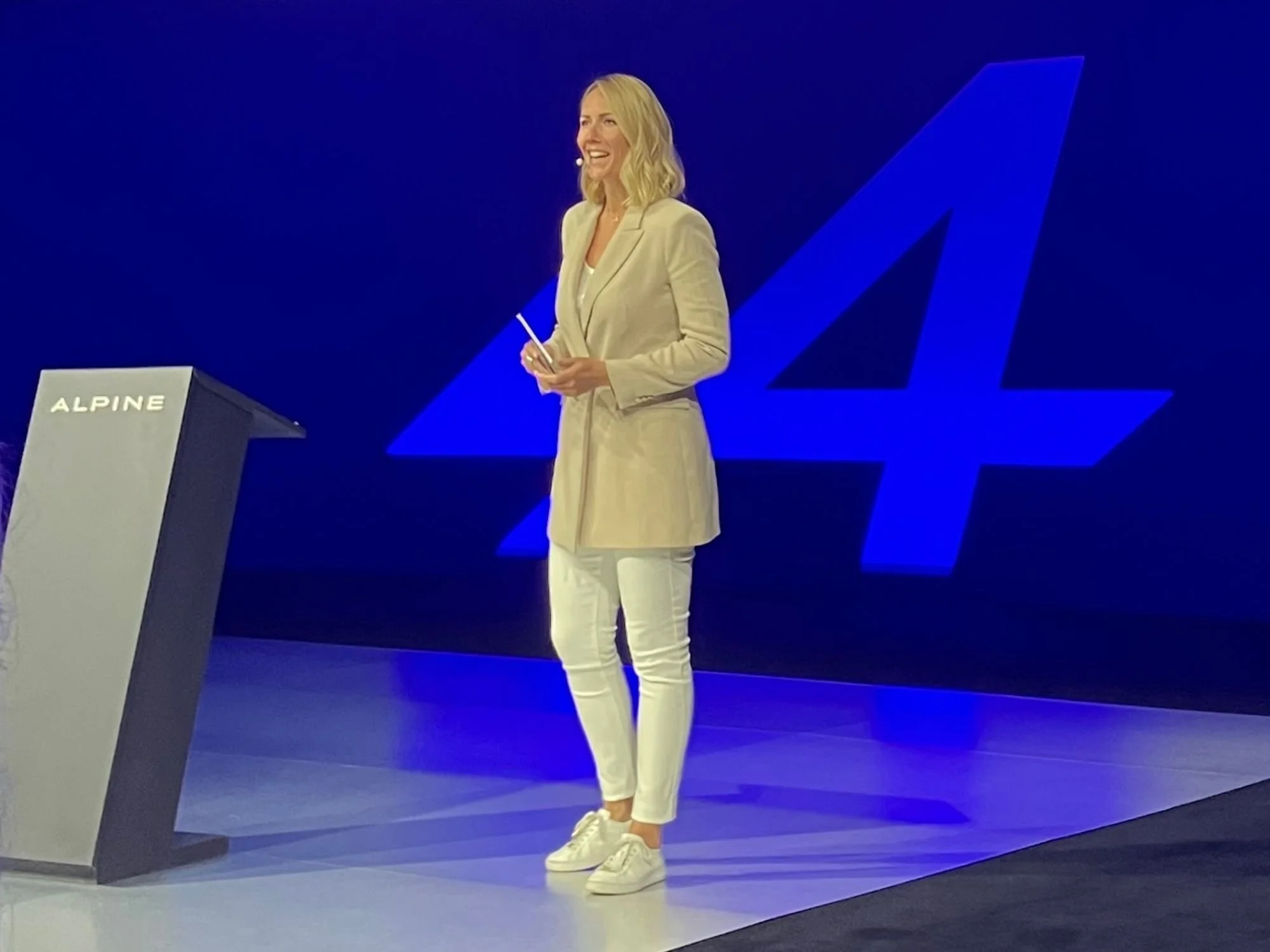 Une femme vêtue d'un blazer beige et d'un pantalon blanc se tient sur une scène avec un fond bleu et un grand logo. Elle sourit et tient une fiche, avec un podium portant l'inscription « ALPINE » à proximité.