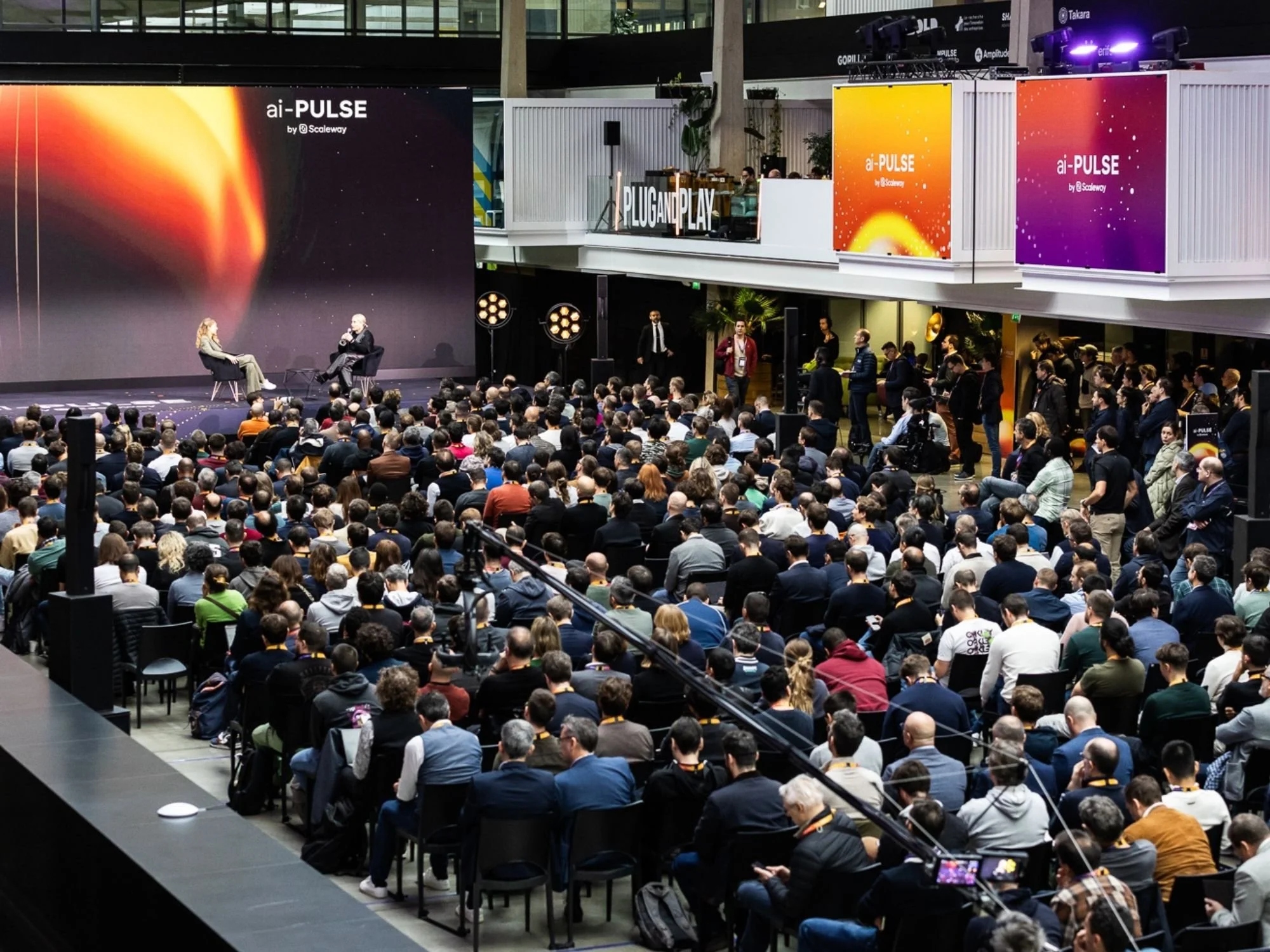 Un large public assis dans une salle de conférence regarde deux personnes discuter sur scène, avec des bannières colorées au-dessus de leurs têtes affichant « ai-PULSE by Scaleway ».