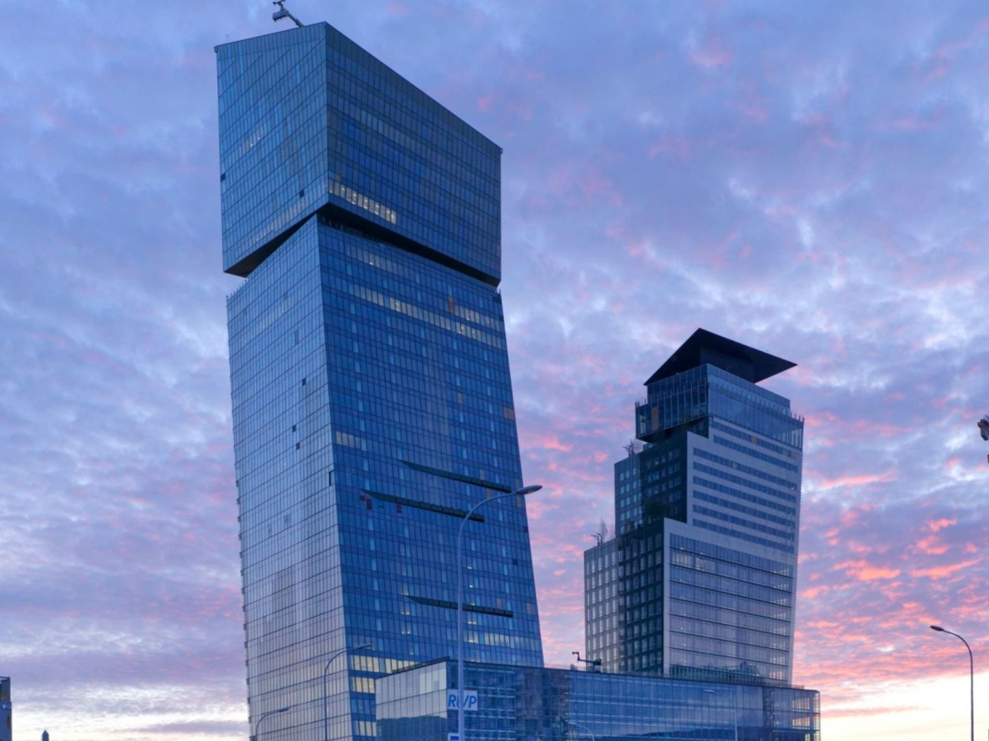 Deux gratte-ciel modernes en verre à l'architecture unique se détachent sur un ciel rose et violet au coucher du soleil.