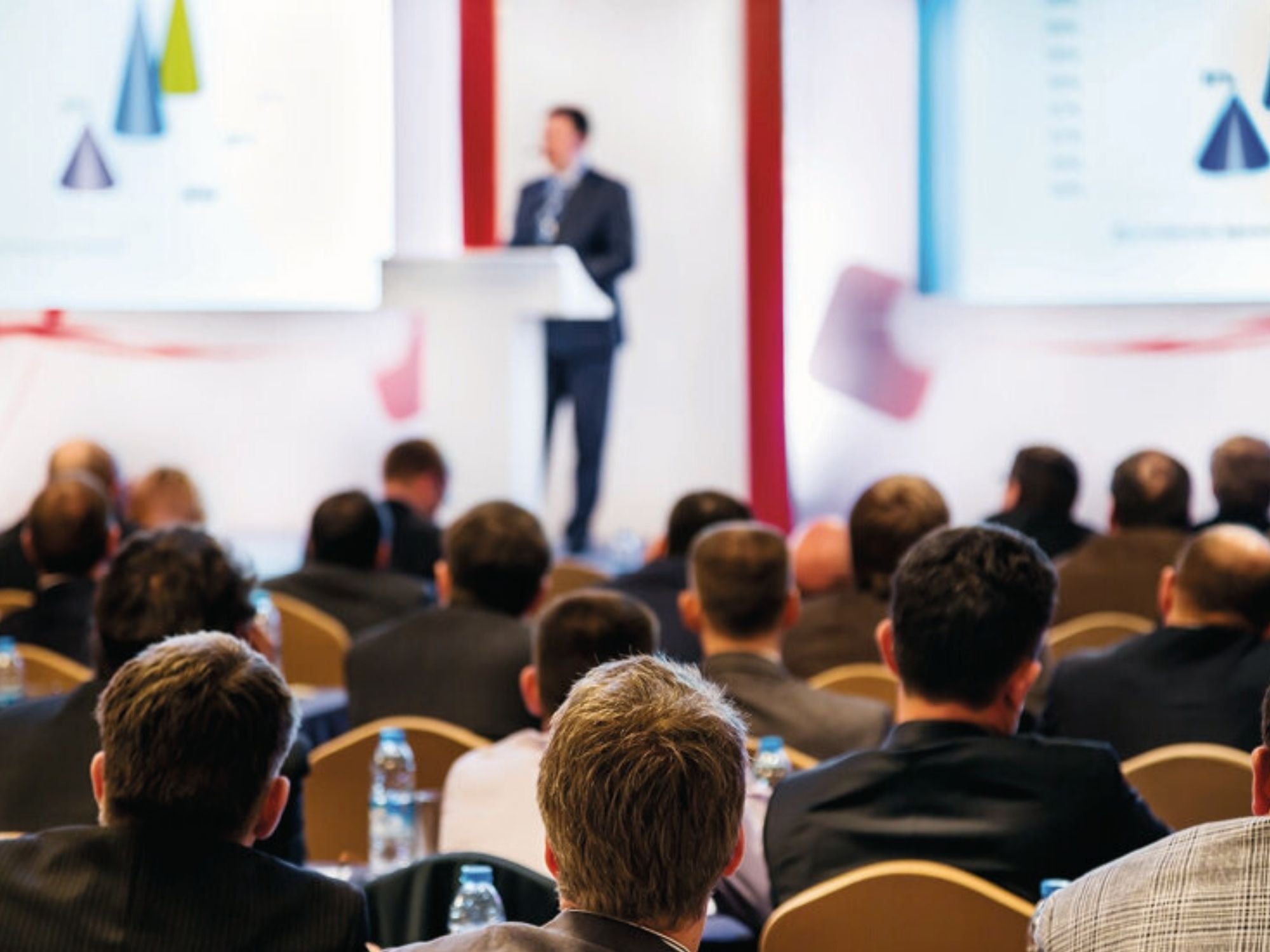Conférence d'affaires avec un public écoutant un orateur sur scène, des diapositives de présentation sur des écrans et des tables avec des bouteilles d'eau.