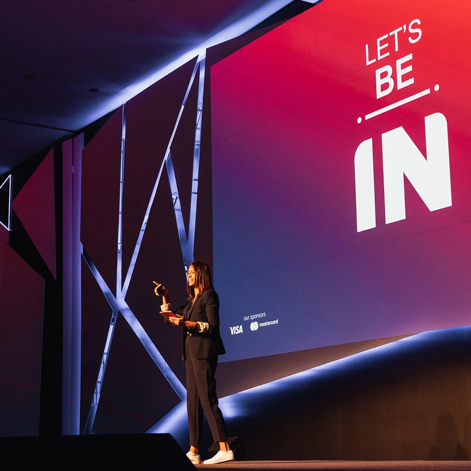 Une femme en tailleur noir fait une présentation sur scène, derrière elle un grand écran affiche la citation motivante « Let's be in » (Soyons présents) et les logos des sponsors Visa et Mastercard.