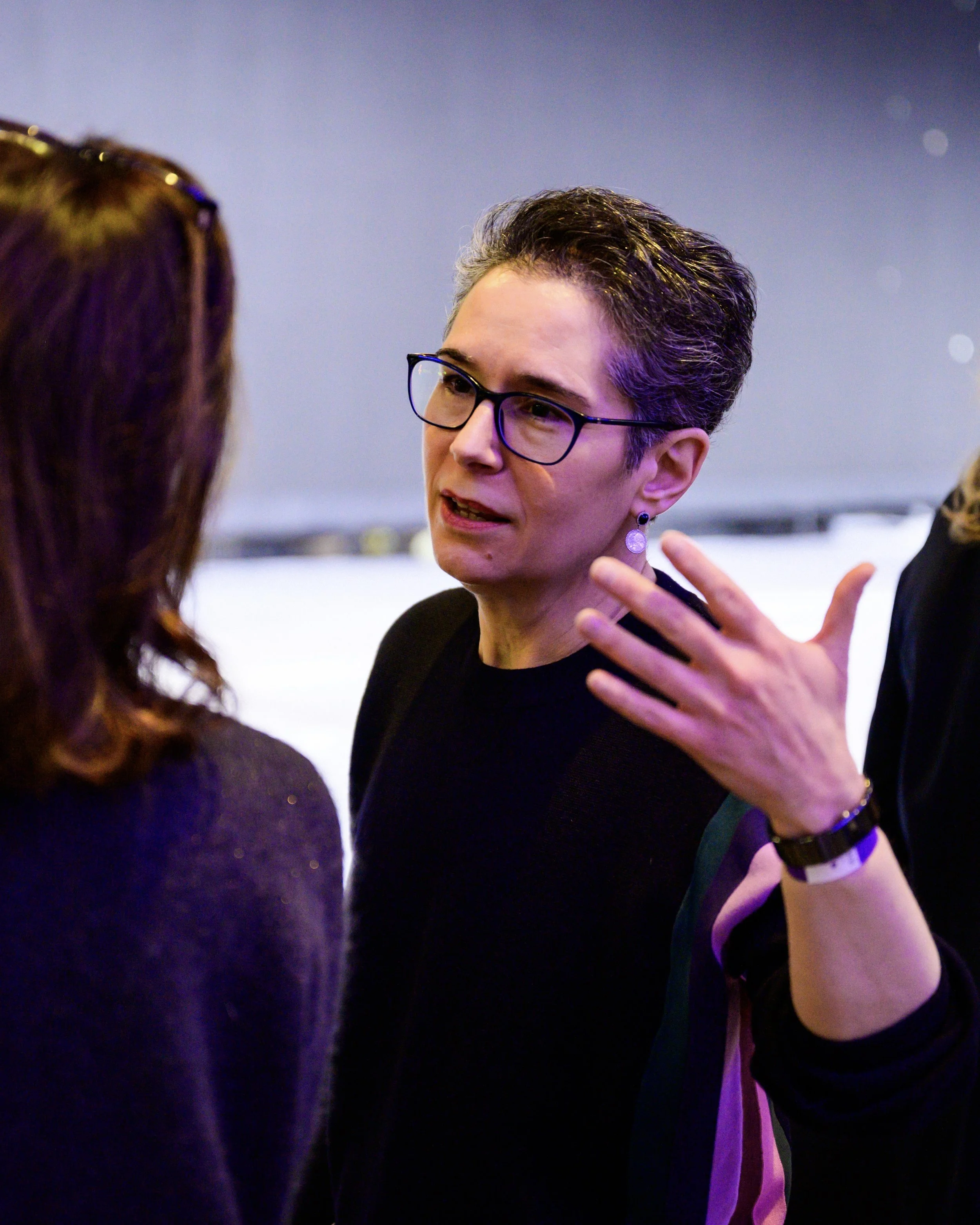 Une femme aux cheveux gris courts, portant des lunettes et des boucles d'oreilles, parle à une autre femme aux cheveux bruns. La femme aux cheveux gris gesticule avec sa main et semble engagée dans une conversation.