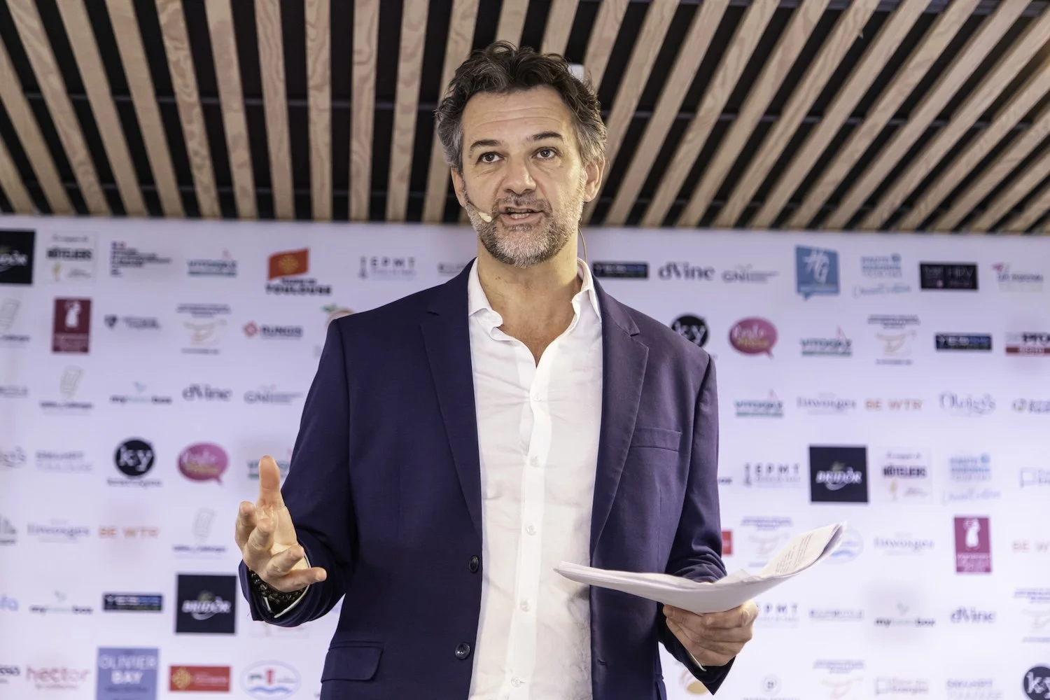 Un homme aux cheveux gris et à la barbe grise, vêtu d'un blazer bleu marine et d'une chemise blanche, s'exprimant lors d'un événement devant un fond comportant divers logos.