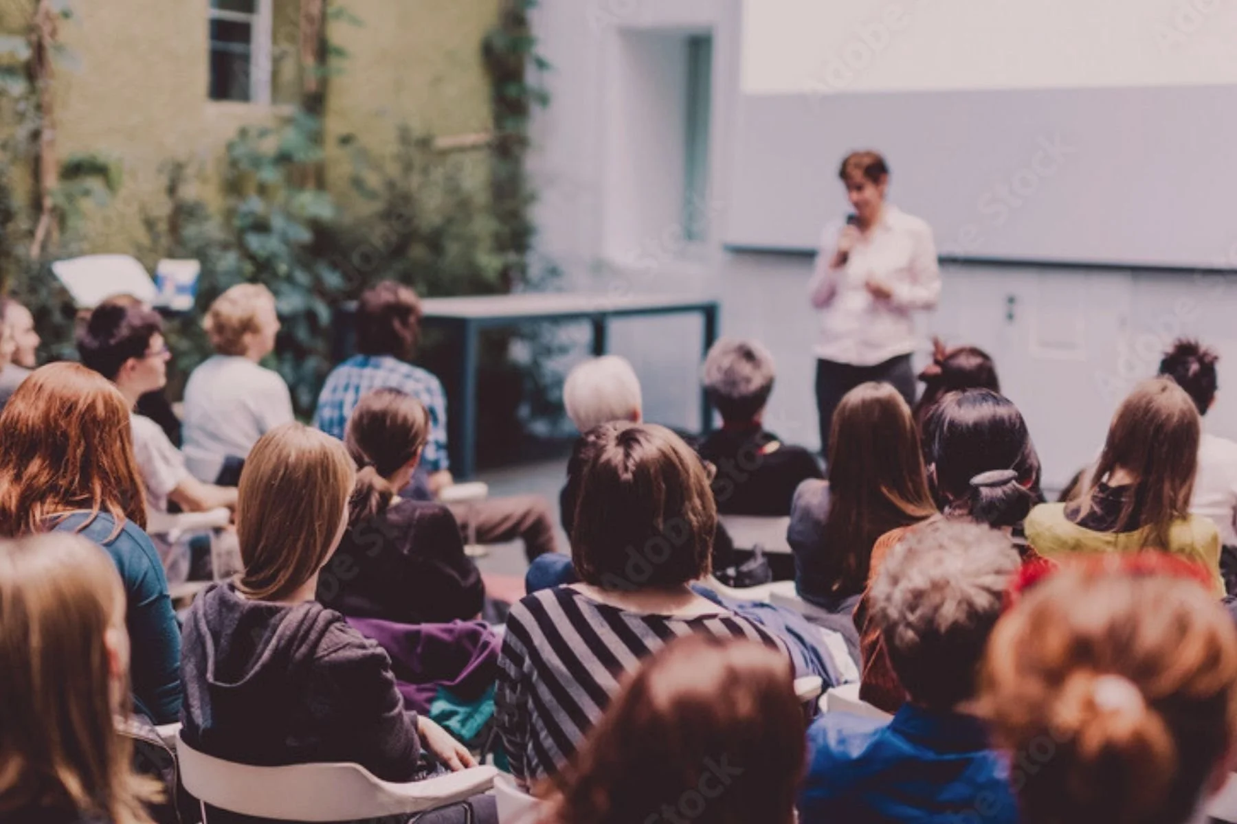 Un groupe de personnes assistant à un séminaire ou à une présentation en plein air, avec une femme qui s'adresse au public près d'une table et d'un écran.
