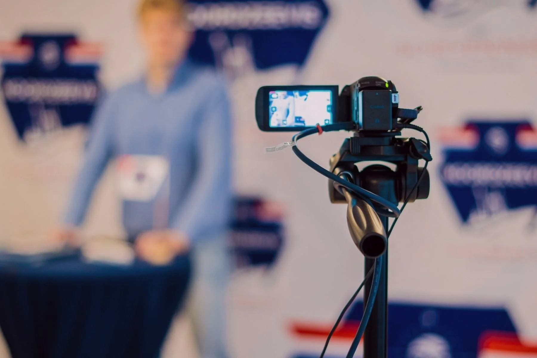 Une caméra sur un trépied filmant une personne à un podium lors d'une conférence de presse ou d'une présentation.