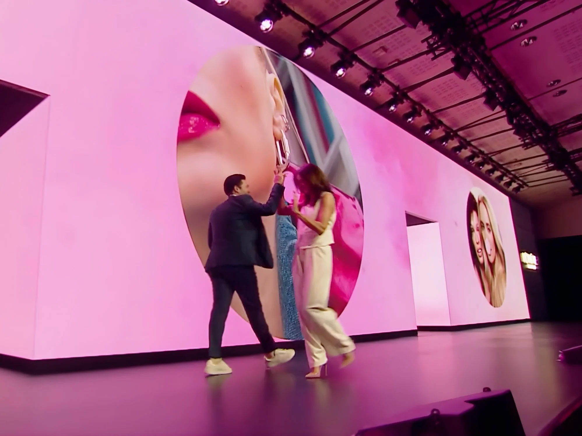 Un homme et une femme sur une scène avec des fonds roses et violets et de grandes images circulaires représentant des lèvres et des femmes, engagés dans une performance ou une présentation.