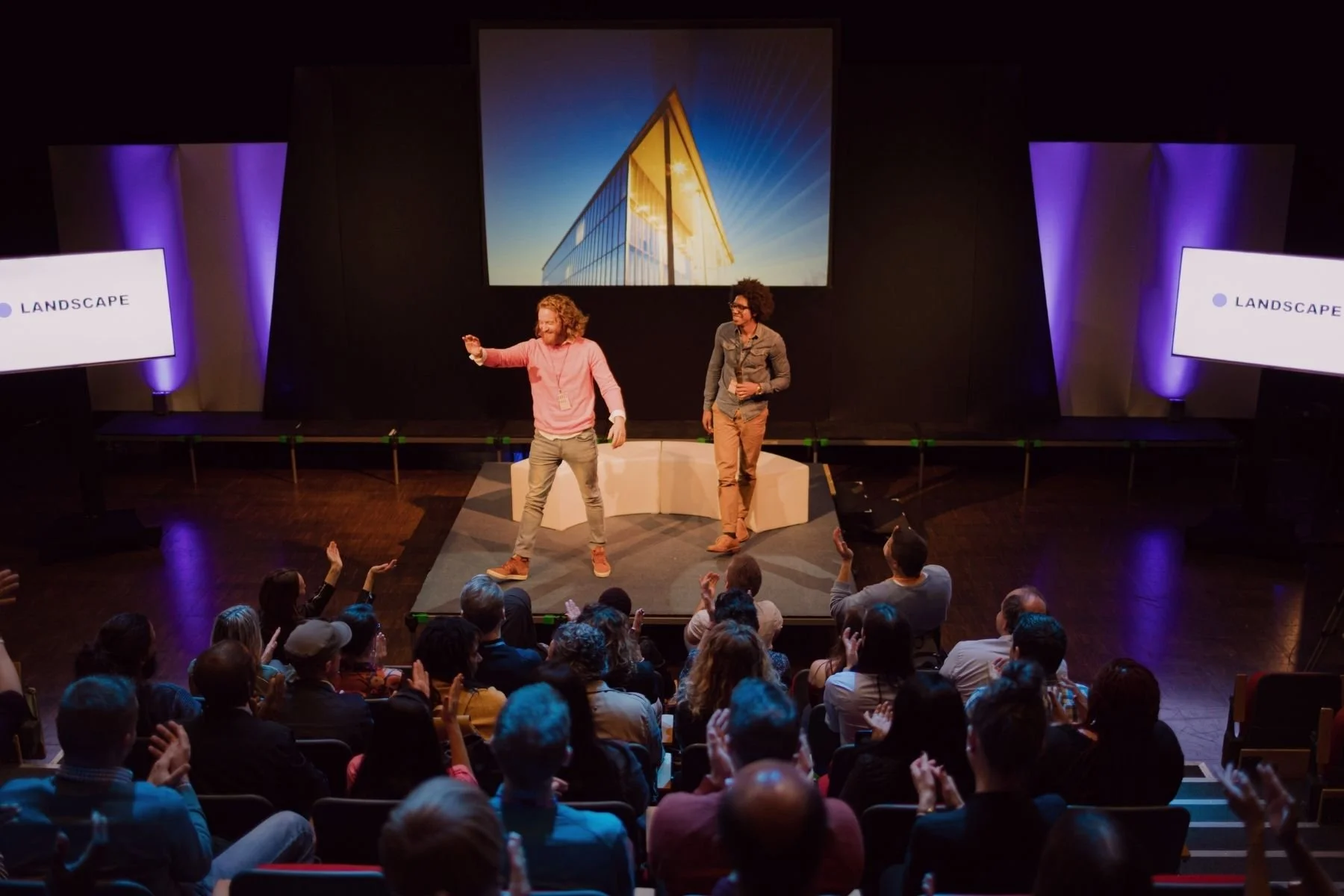 Deux présentateurs sur scène lors d'une conférence ou d'un événement, avec le public qui applaudit et lève les mains. La scène est équipée de grands écrans affichant le mot « LANDSCAPE » et d'une toile de fond représentant un bâtiment moderne baigné de soleil.