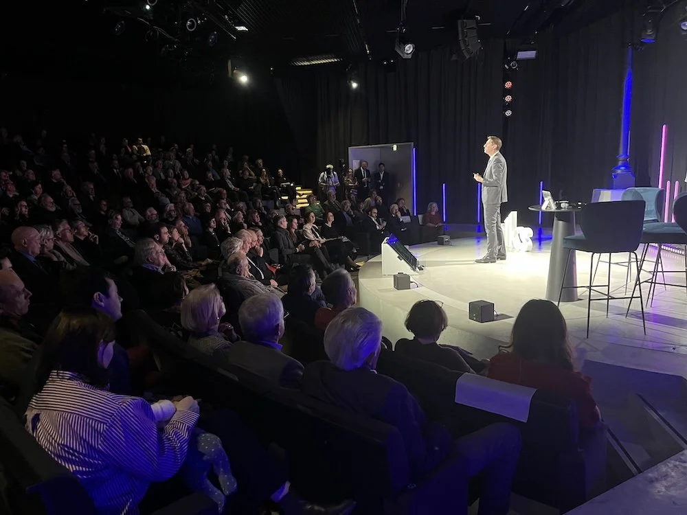Un homme en costume gris fait une présentation sur scène devant un public dans un auditorium sombre. La scène est équipée d'une petite table et de chaises, et il y a des lumières et des écrans à l'arrière-plan.