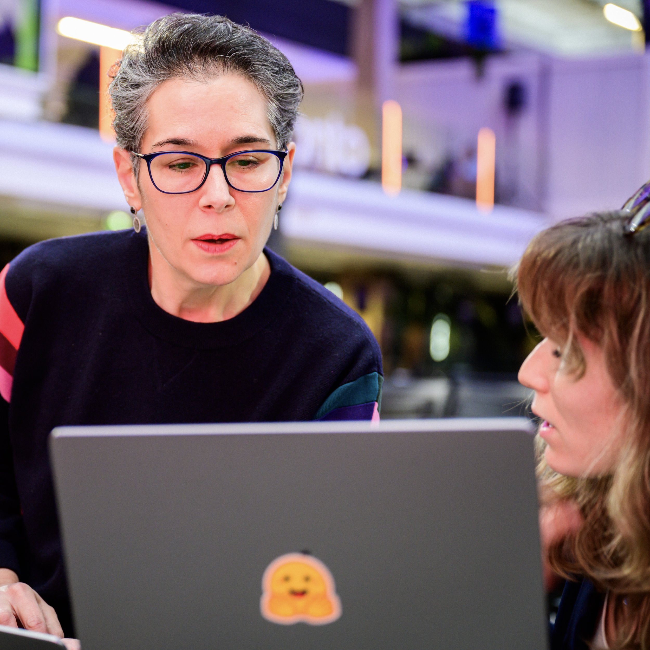 Deux femmes discutent de leur travail à une table avec un ordinateur portable dans un bureau intérieur bien éclairé ou un environnement de travail technologique.