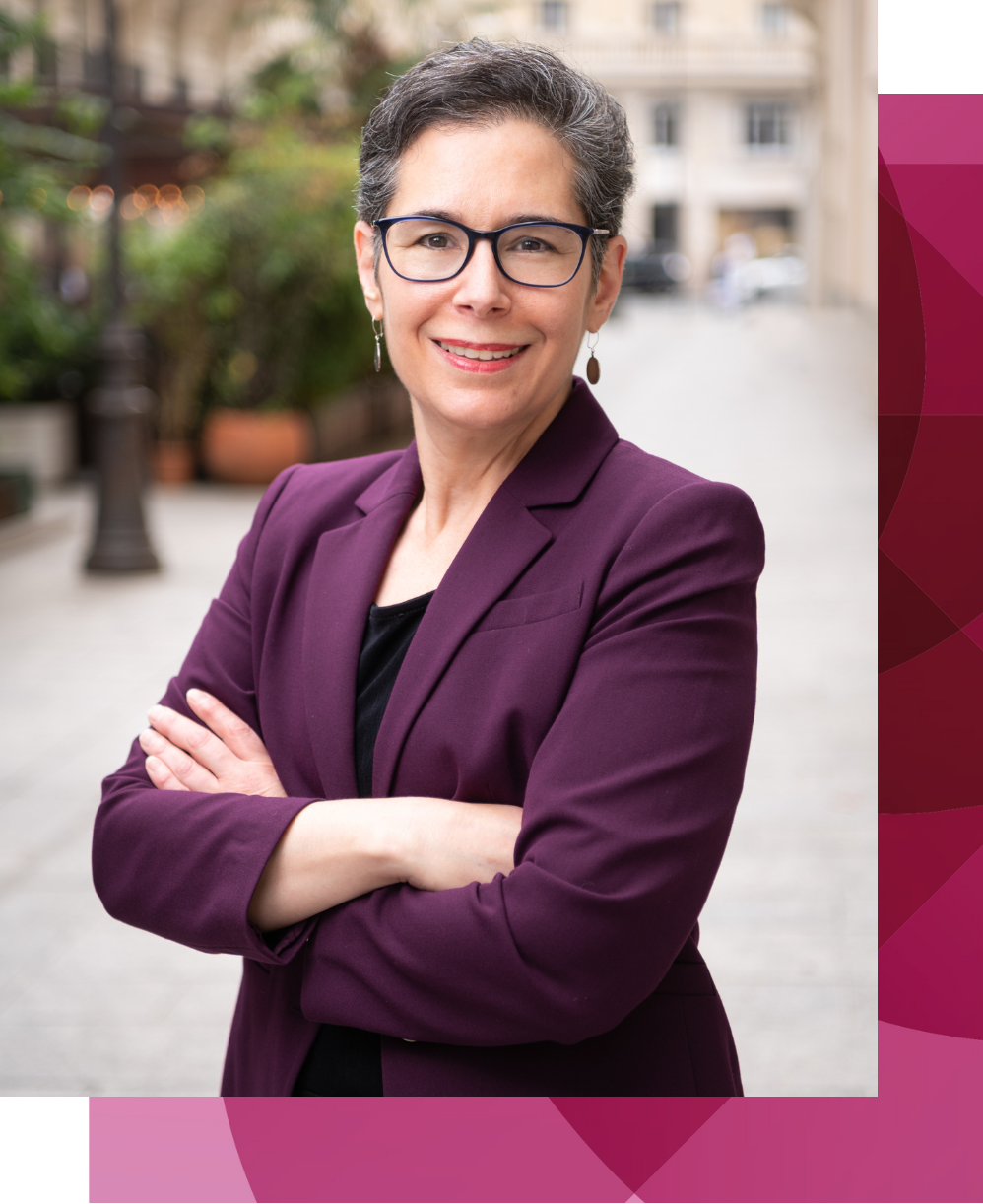 Une femme professionnelle aux cheveux gris courts, portant des lunettes et des boucles d'oreilles, vêtue d'un blazer violet et d'un haut noir, debout à l'extérieur, les bras croisés, souriante et face à la caméra.