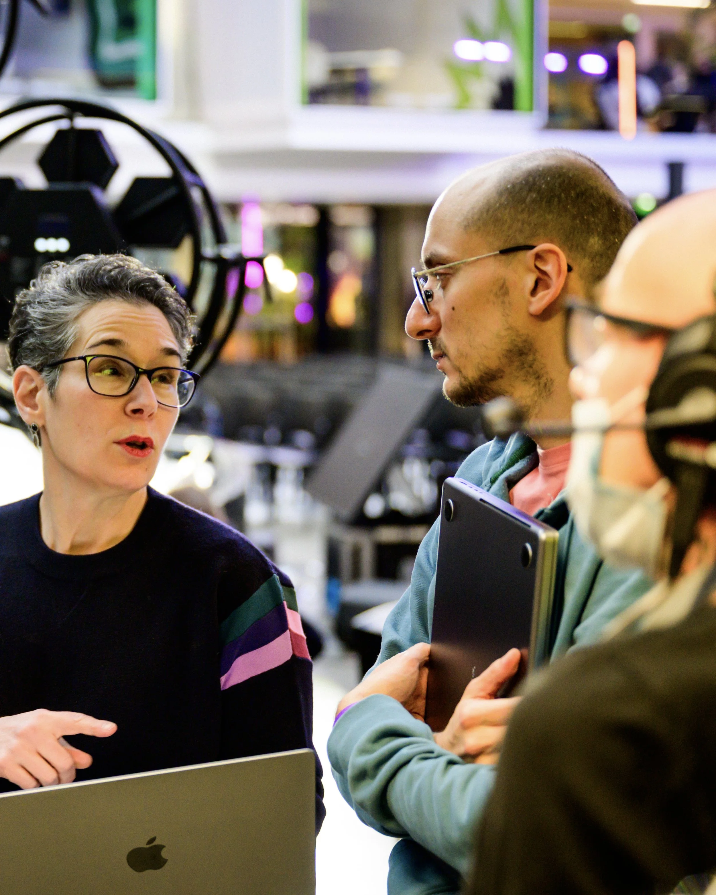 Trois personnes discutent dans un magasin d'électronique, avec des ordinateurs portables et du matériel électronique visibles à l'arrière-plan.