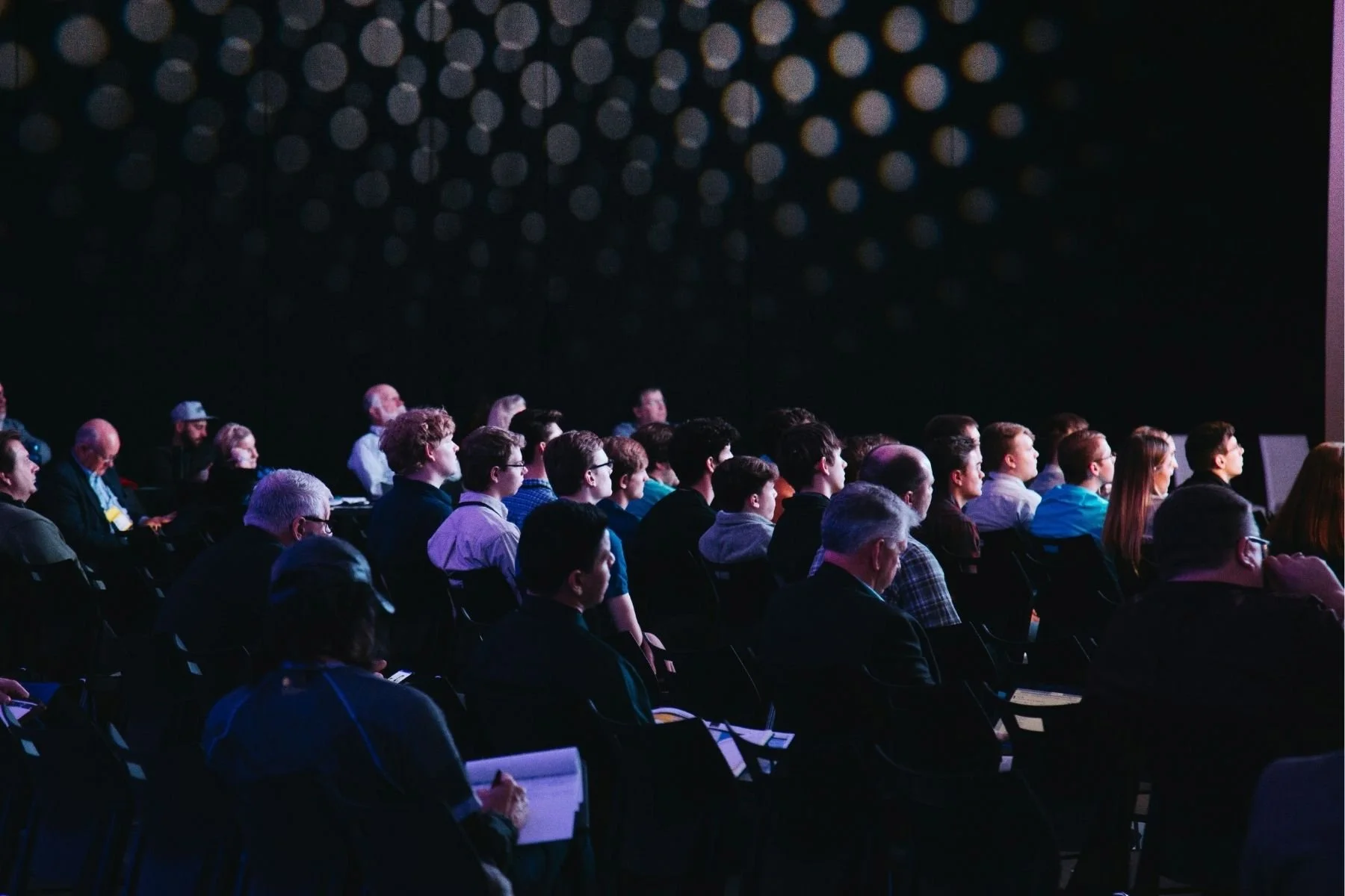 Un public composé de personnes diverses, assises et regardant attentivement une présentation ou un spectacle dans une salle obscure.