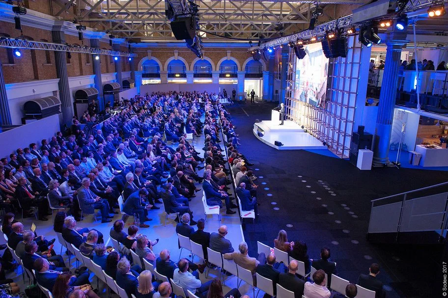 Un large public assis dans un auditorium couvert regarde un orateur sur scène, avec un grand écran, un éclairage bleu et des fenêtres en arc décoratives.