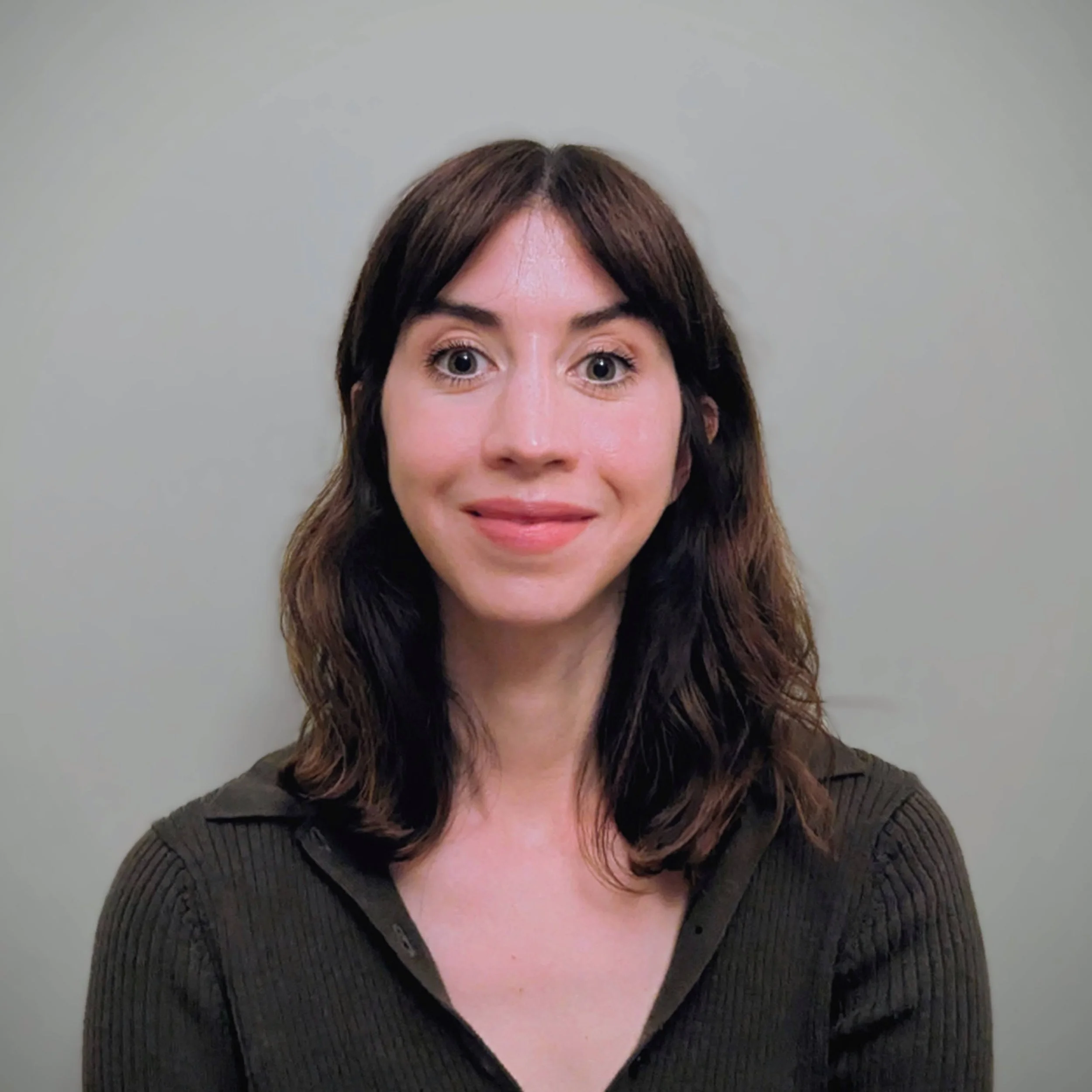 Portrait of a woman with shoulder-length brown hair, wearing a dark green ribbed shirt, smiling against a plain light gray background.