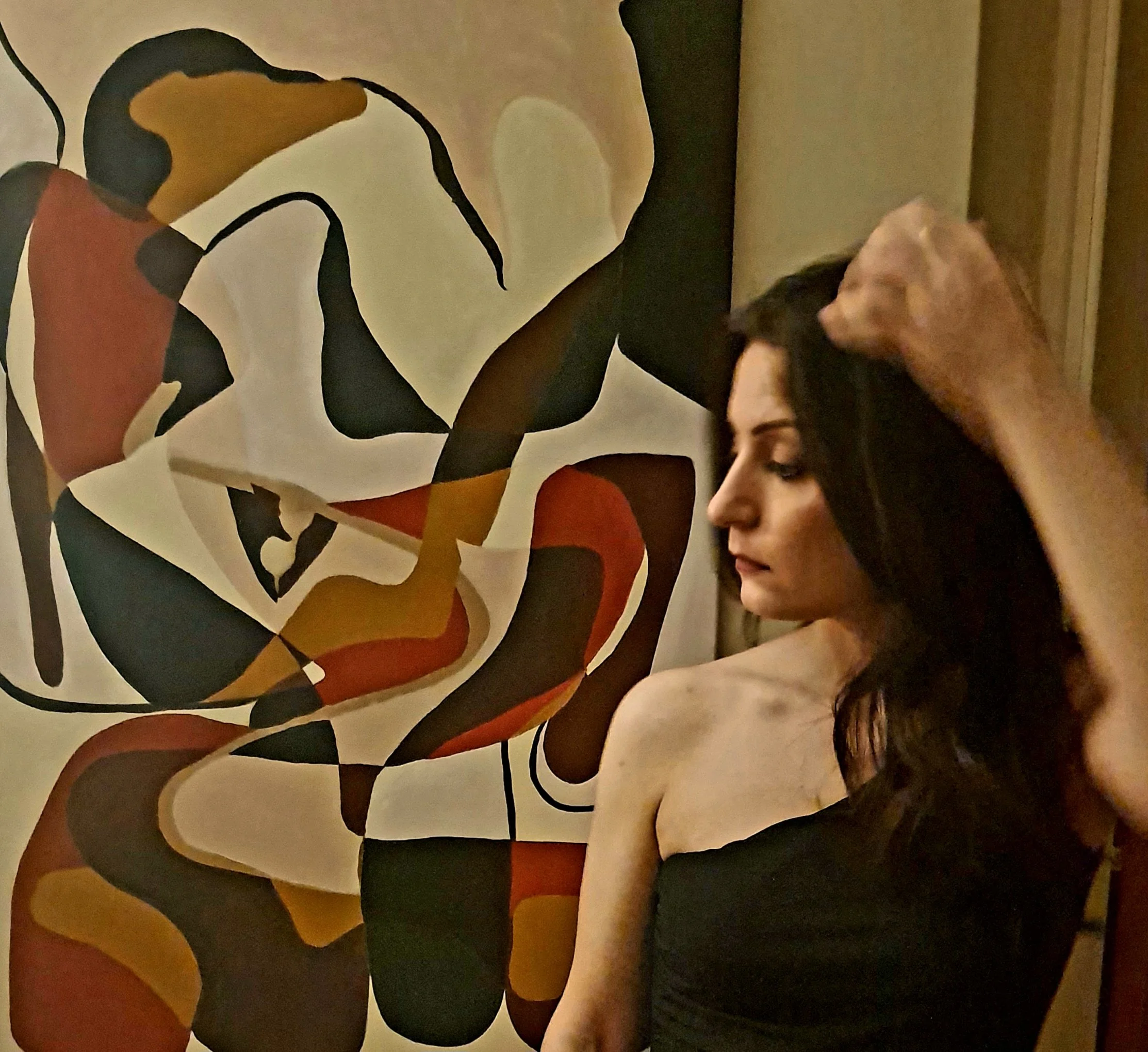 A woman sitting on a black stage floor, with her legs crossed, dressed in dark clothing, with two bottles, a circular object, and a stone behind her, against a plain gray background.