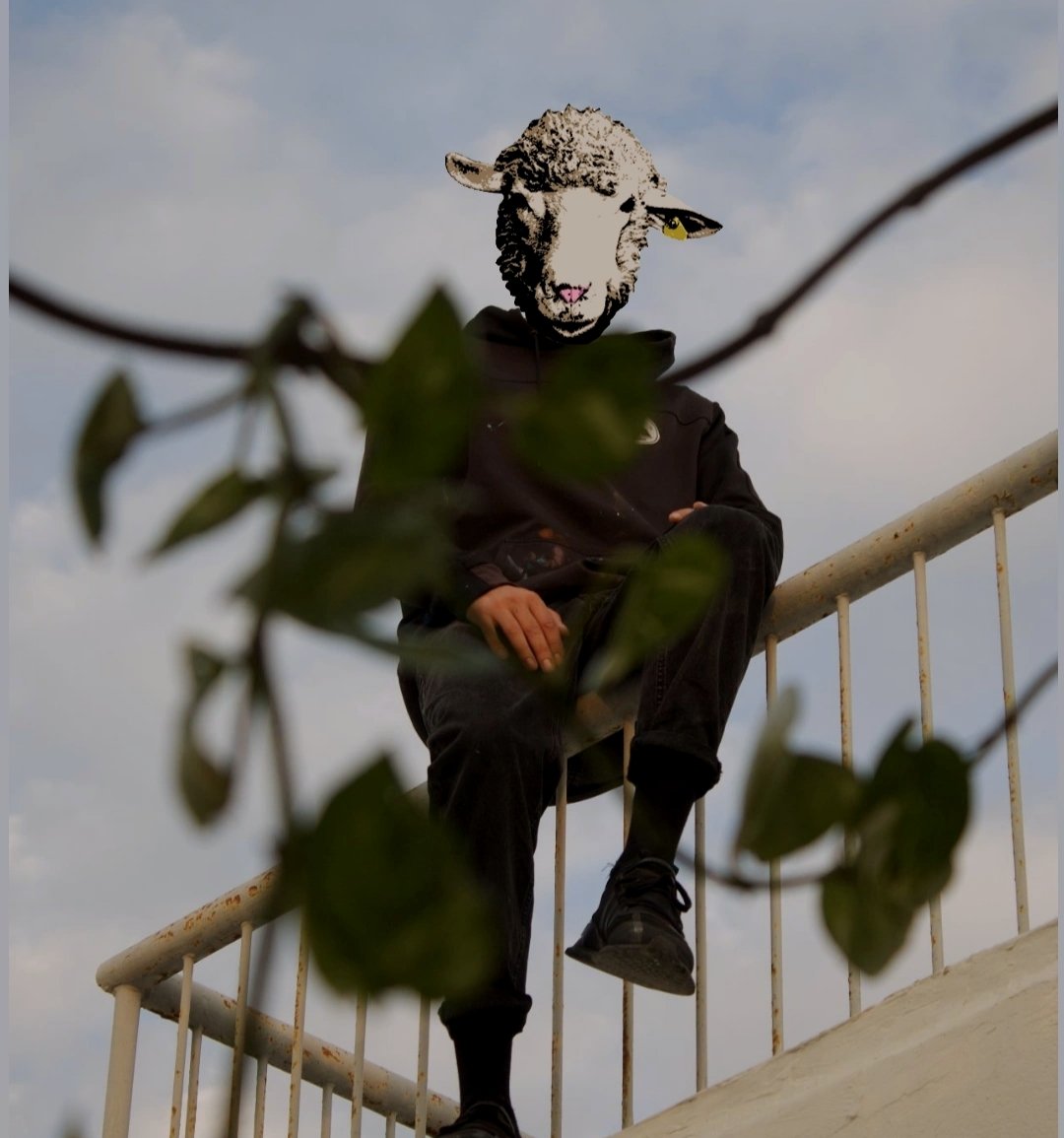 A woman sitting on a black stage floor, with her legs crossed, dressed in dark clothing, with two bottles, a circular object, and a stone behind her, against a plain gray background.