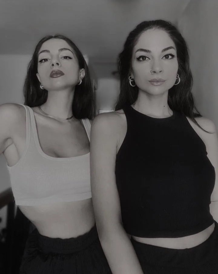 A woman sitting on a black stage floor, with her legs crossed, dressed in dark clothing, with two bottles, a circular object, and a stone behind her, against a plain gray background.